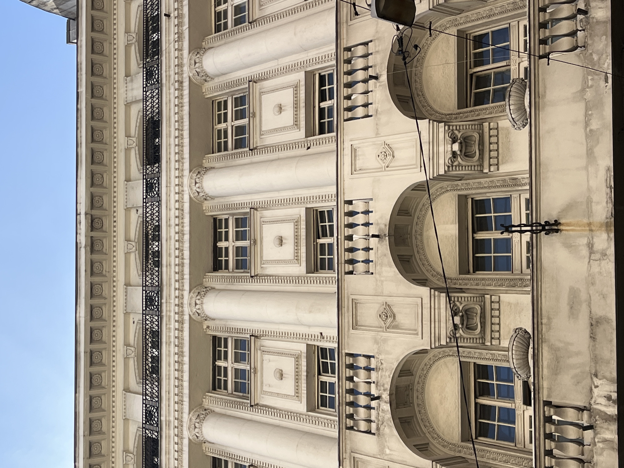 The image shows a multi-story building in Belgrade, Serbia. The architecture exhibits elaborate facades with decorative moldings, columns, arched window frames, and wrought-iron balconies. The building’s exterior is light-colored, and the sky is visible in the background. There are no people visible in the image.