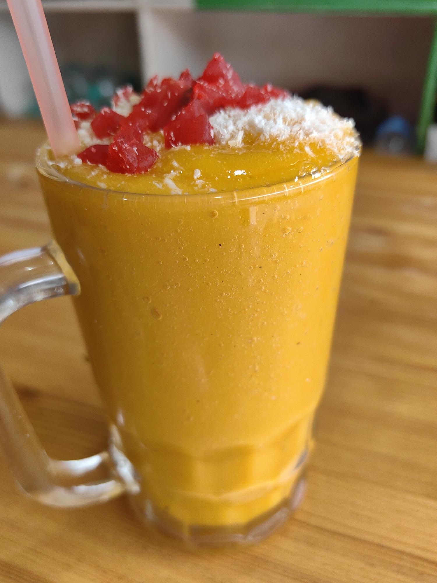 A tall, clear glass mug filled with a vibrant orange smoothie sits on a wooden table. A pink straw is inserted into the smoothie, which is topped with shredded white material, possibly coconut, and small pieces of bright red candied fruit. The mug has a clear handle attached to its side. The background is blurred, showing hints of shelving and muted colors, suggesting an indoor setting, possibly a cafe or shop. The scene is well-lit, highlighting the texture and color of the drink. The image appears to be a close-up shot focusing on the beverage. This image was taken in Vashisht, India.