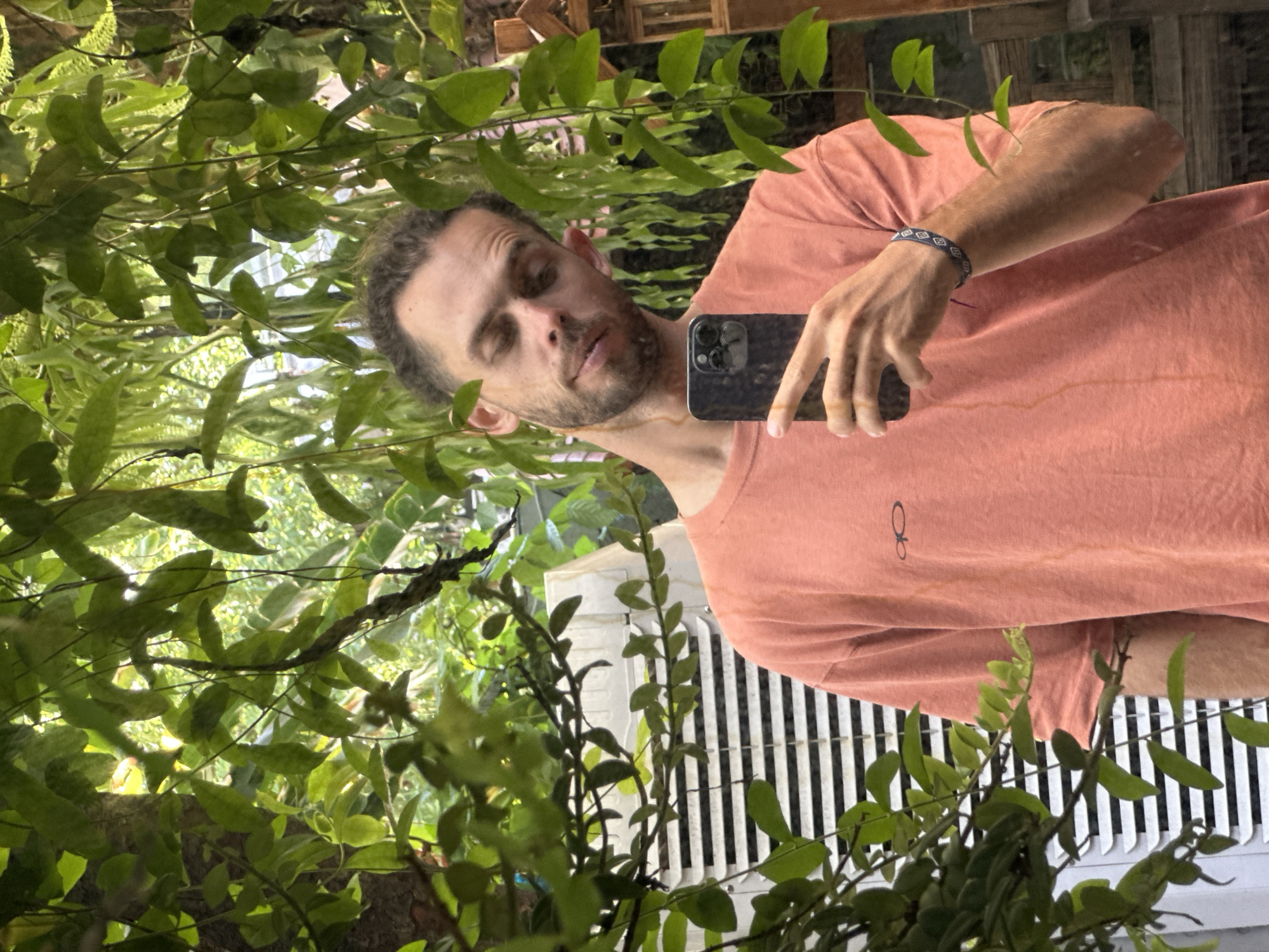 A man with short, curly, light brown hair and stubble is taking a mirror selfie outdoors. He is wearing a coral-colored t-shirt with a small black logo on the left chest. He has a bracelet on his left wrist with a geometric pattern. The background is lush with green foliage, with vines hanging down and obscuring parts of the scene. To the left of the man, a white air conditioner unit with horizontal slats is visible. The lighting suggests it is daytime, with sunlight filtering through the leaves. The setting appears to be a residential area or garden.