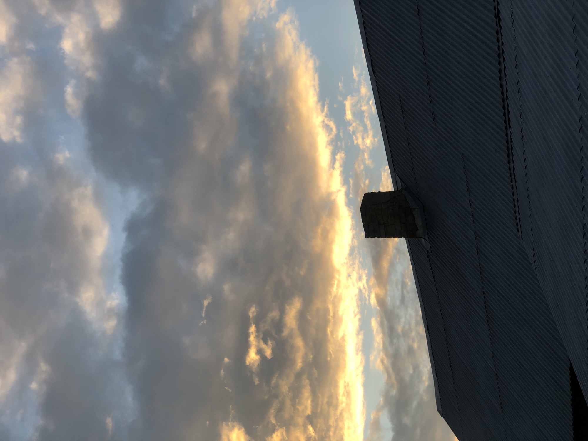 This image depicts a cloudy sky at sunset, with the sun's rays illuminating the clouds with a warm, golden hue. The foreground is dominated by the dark, corrugated metal roof of a building, angled from the bottom right to the top left of the frame. A small, rectangular stone structure protrudes from the roof, possibly a chimney or ventilation shaft. The sky is filled with a mix of dark, stormy-looking clouds and lighter, wispy clouds tinged with orange and yellow from the setting sun. The overall mood is dramatic and somewhat somber, with a hint of the beauty of nature. The time of day is clearly sunset, given the lighting and the colors of the clouds. There is no visible text in the image. The location is identified as Zhukovo, Russia, which might suggest a rural or industrial setting, consistent with the building's roof. The activity is simply the observation of the sunset and the sky.