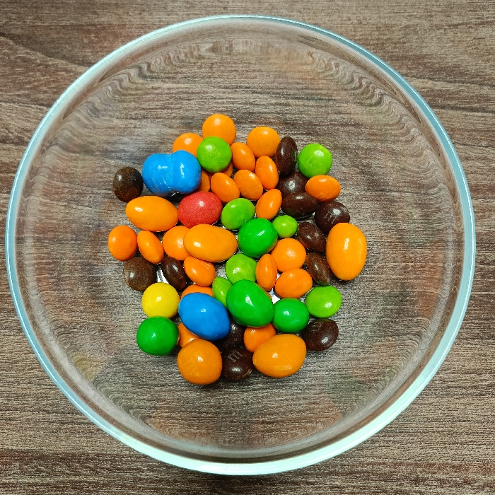 A clear glass bowl contains a collection of candy-coated chocolates, consisting of both ovular and spherical forms. The color palette comprises bright orange, lime green, dark brown, light blue, yellow, and red candies. Some dark brown candies feature a visible 'm' insignia. The bowl is positioned on a light brown wooden surface displaying a distinct grain pattern. No information is available within the image to determine the geographical location of Markovskii, Russia.