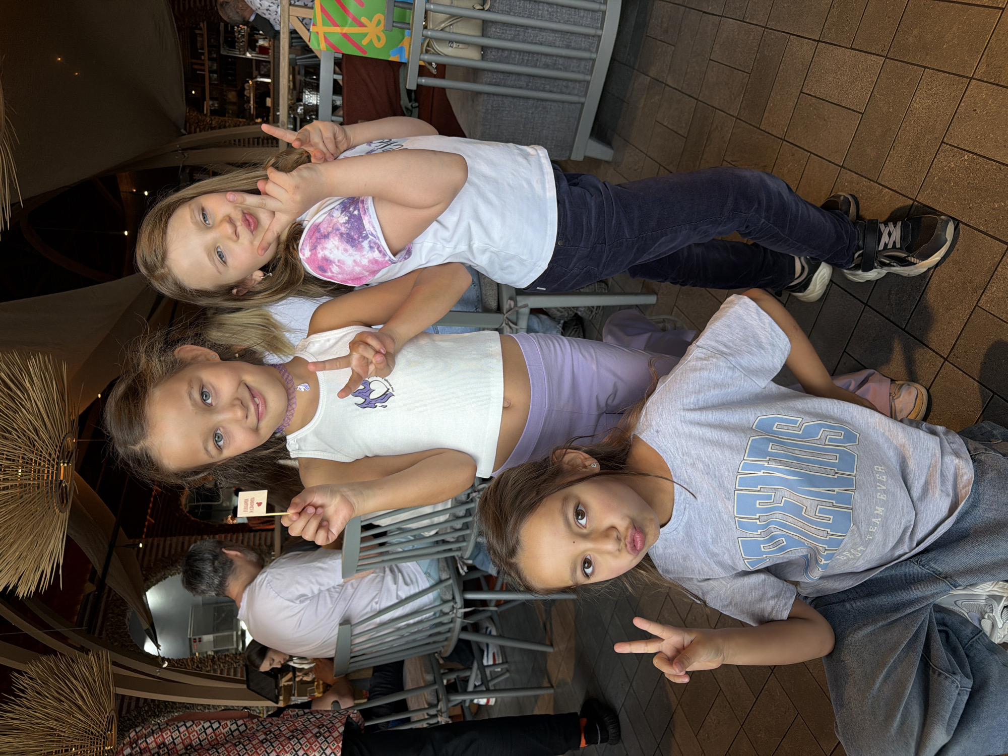 Three young girls are seated at a table. The girl on top is wearing a white t-shirt with a purple/pink design on the sleeves and dark pants. The middle girl wears a white tank top, purple pants, and a purple necklace. She is holding a small sign on a stick. The girl on the bottom is wearing a light blue t-shirt and jeans. All three girls are making peace signs with their hands and smiling at the camera. They are in an indoor dining establishment, with other patrons visible in the background. The location cannot be determined from the image, including the city of Krasnoyarsk, Russia.