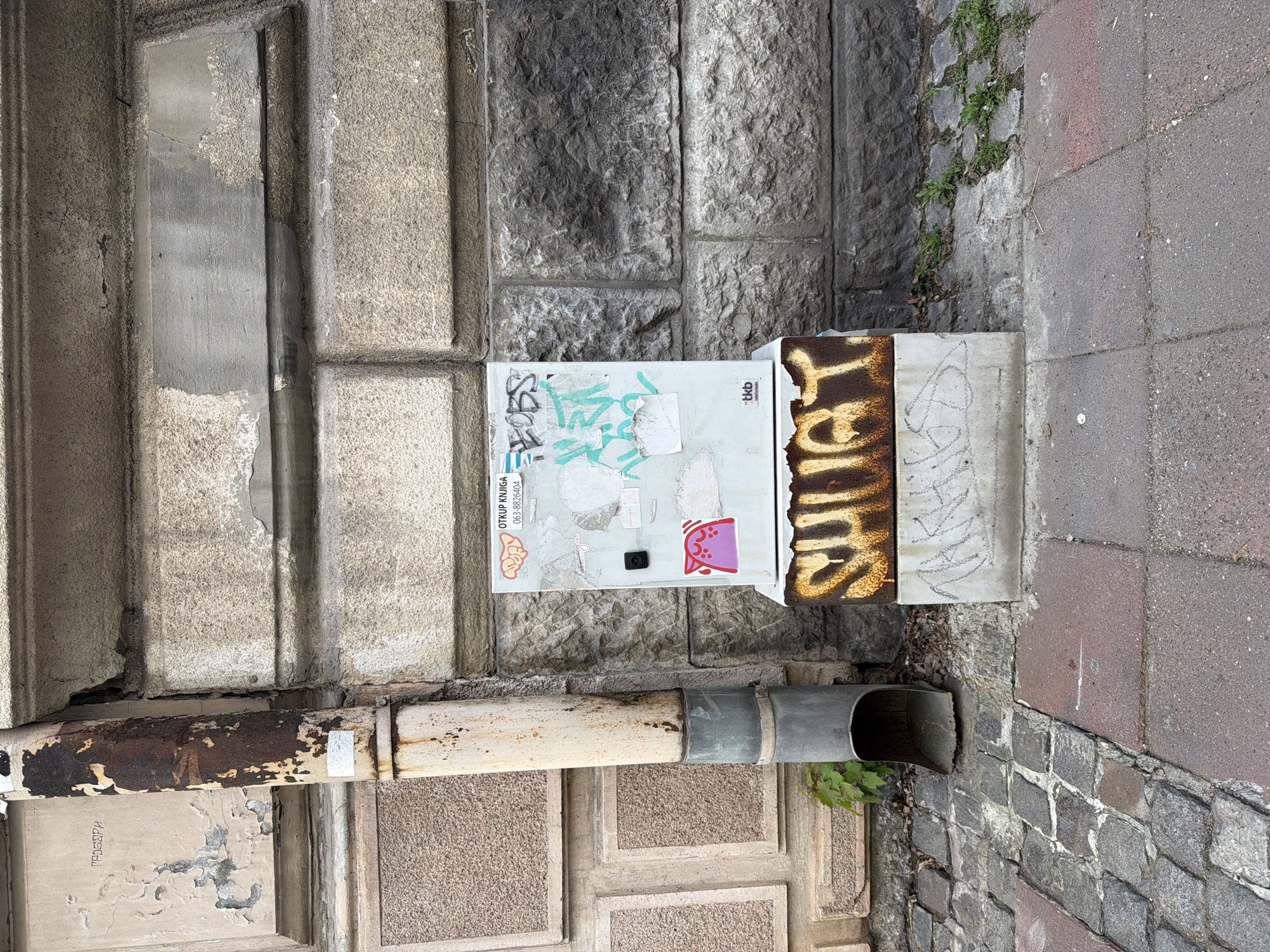 A ground-level, eye-level shot depicts a section of an urban landscape in Belgrade, Serbia. A light gray electrical box is mounted on a stone wall, covered in graffiti, stickers, and a large rusty brown marking, oriented vertically. A metal downspout runs horizontally below the box, progressing from rusty to gray, terminating with a flared outlet near the cobblestone street. The wall behind the box and above the downspout is constructed of large, textured gray stones. To the left, a concrete wall with a grooved texture is visible. To the right, there are reddish-brown pavement tiles bordering the cobblestone street. Sparse green vegetation grows between the stone wall and the pavement.