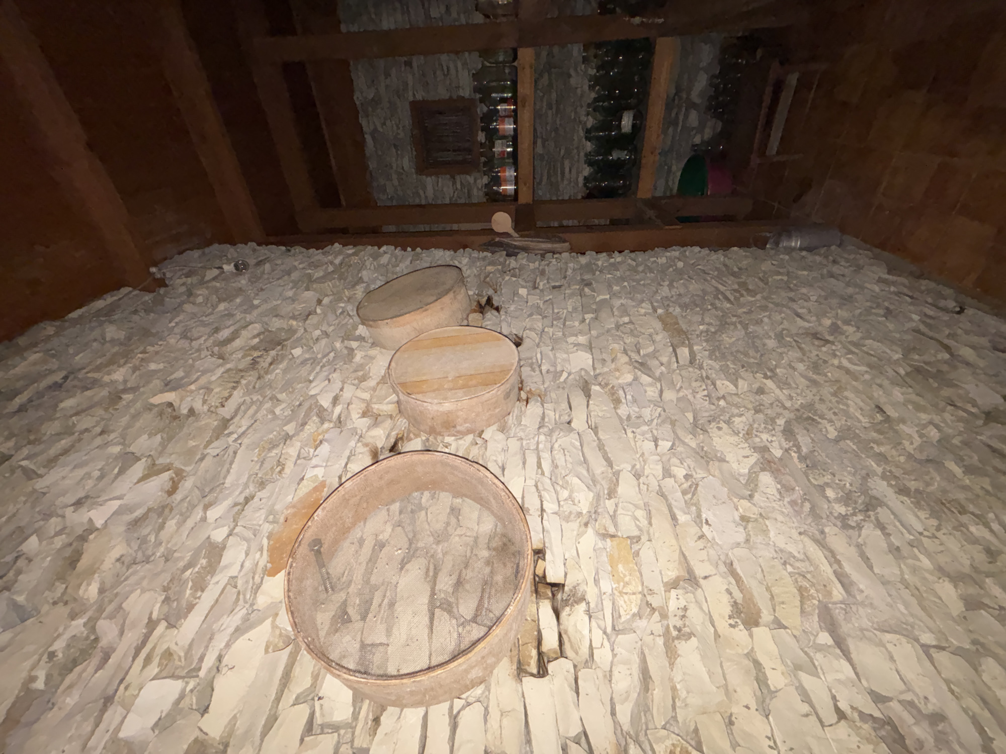 The image shows the interior of a rustic room, likely a cellar or storage area, with a stone wall and wooden ceiling beams. On the stone wall, three circular sieves are hung. The largest sieve is on the left, with a metal mesh and wooden frame. To its right are two smaller sieves, also with wooden frames. The room has shelves made of wood on the right side, filled with glass bottles, some appearing to contain liquids, and what seem to be jars. A small wooden stool and a few buckets are also visible on the floor. The lighting is dim, suggesting an underground or dimly lit space. The overall impression is one of storage or preparation of food or beverages, common in rural or historical settings. The specific location in Gudauta, Abkhazia, suggests a historical or traditional building, possibly a winery or food storage cellar given the presence of bottles and sieves.