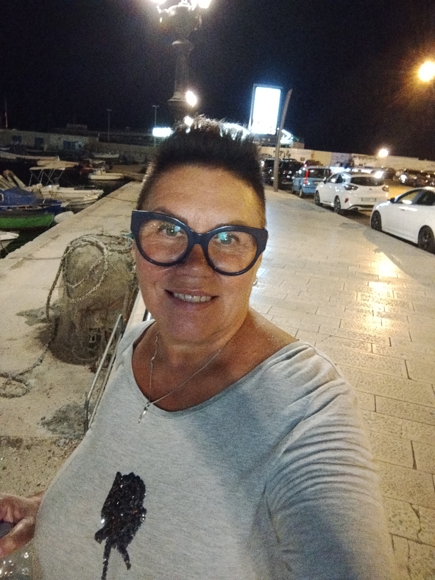 The image captures a woman with short, dark hair and thick-rimmed glasses, taking a selfie at night in the city of Bari, Italy. She is wearing a light gray t-shirt with a dark, sequined figure on the front and a silver necklace. The background reveals a waterfront scene with several boats docked. To the right, a paved walkway lined with parked cars is visible, illuminated by streetlights. The overall atmosphere is casual and urban, typical of a seaside promenade at night.