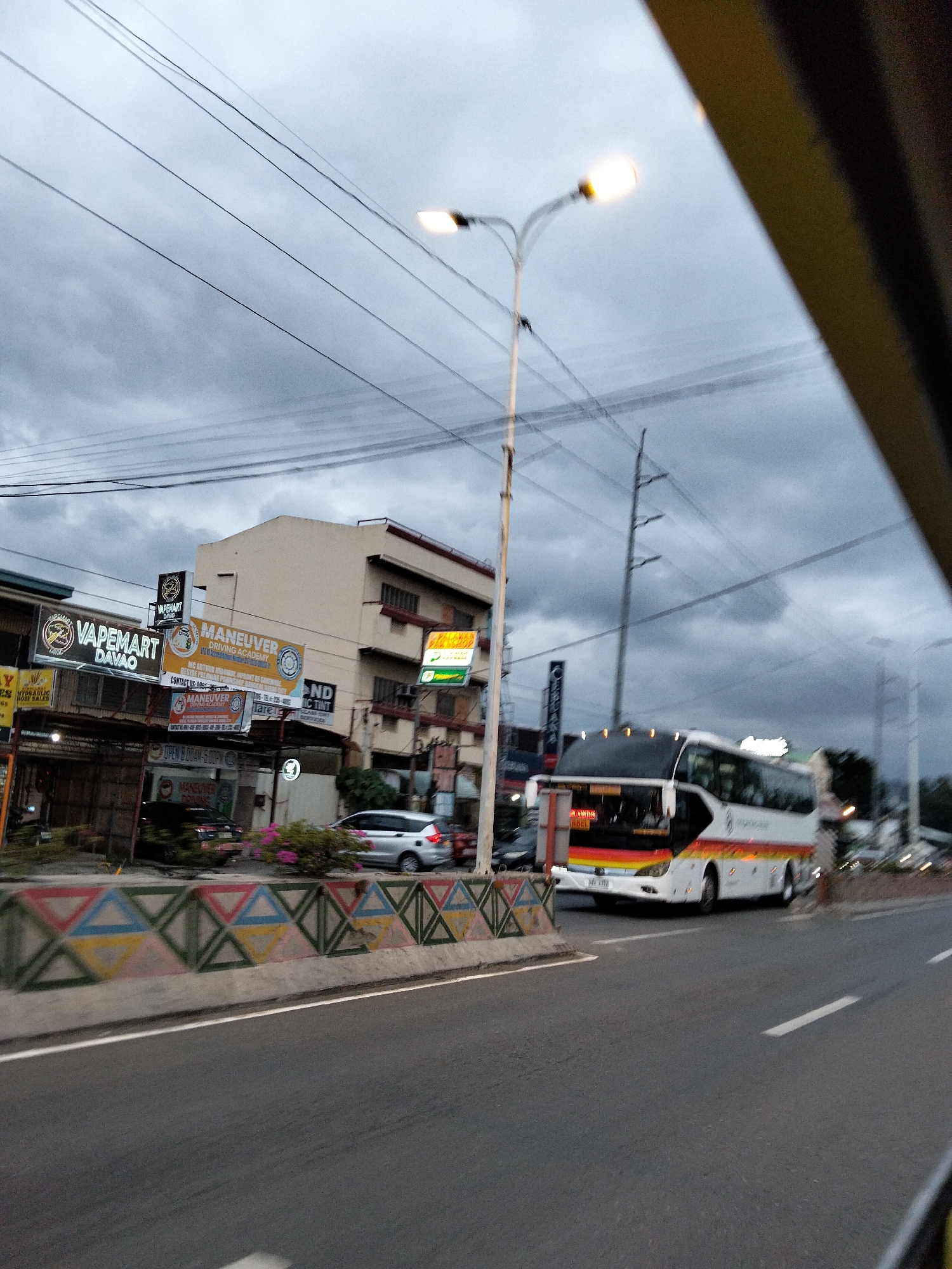 The image shows a wide shot of a street in Davao City, Philippines, on an overcast day. A white intercity bus with red, orange, and yellow stripes along the side is traveling on the right side of the road. Several buildings line the left side of the street, including one with a sign that reads "VAPEMART DAVAO" and another displaying "MANEUVER DRIVING ACADEMY." A street light is visible in the upper left quadrant of the image. A concrete barrier with a colorful triangular pattern separates the road from the sidewalk. The sky is filled with dark, heavy clouds, suggesting an impending rain shower. The overall scene depicts a typical urban environment in Davao City, with commercial establishments and public transportation operating under a cloudy sky.