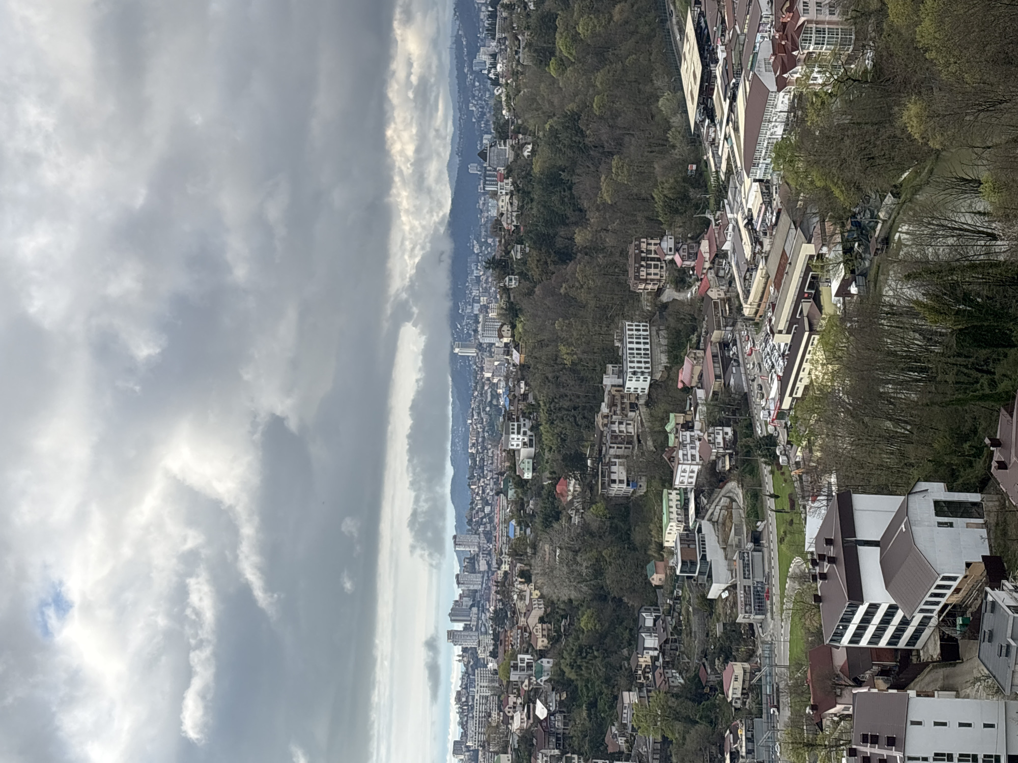 The image shows an elevated view of Sochi, Russia, likely taken from a higher vantage point in the city. The sky is overcast with dense, gray clouds. In the distance, a range of mountains can be seen, partially obscured by haze. The city itself consists of densely packed buildings and residential structures, interspersed with areas of trees. In the foreground are more buildings, some with distinctive rooflines, and roads that wind through the urban landscape. No people are discernible in the image.