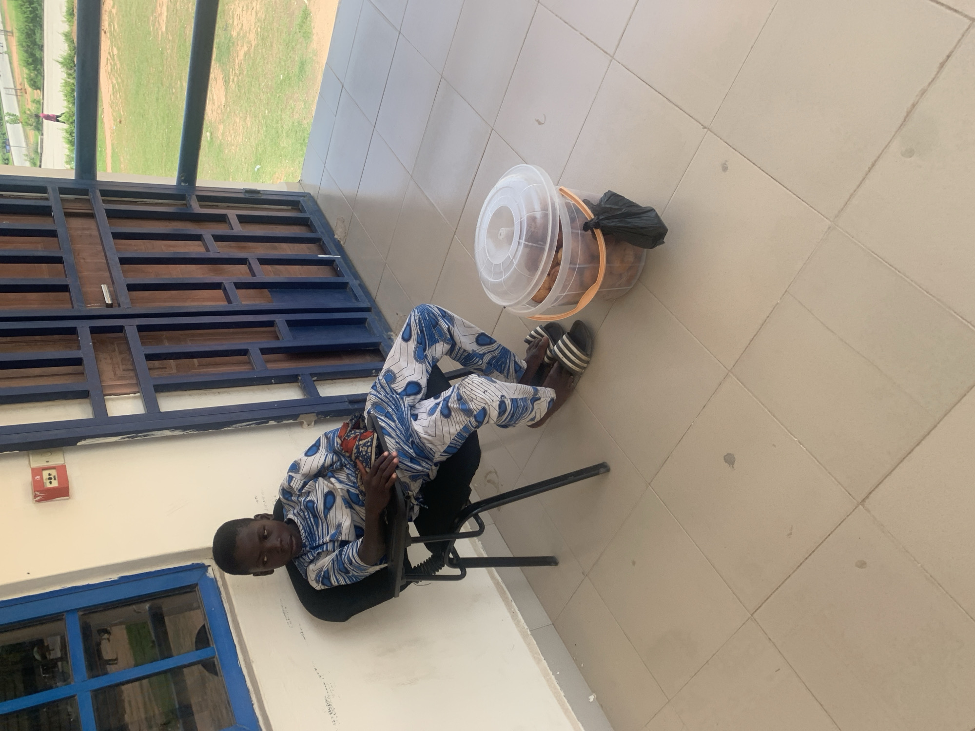 The image shows a young boy sitting on a black metal chair on a tiled outdoor patio. He is wearing a blue and white patterned shirt and black pants, with black and white striped sandals on his feet. The boy is leaning back in the chair and looking towards the camera. To his right, a clear plastic bucket with an orange handle is filled with what appears to be fried food items, possibly plantains, and a black plastic bag is tied to the handle.
The setting appears to be a walkway or veranda, with a tiled floor and a light-colored wall. A blue railing with wooden slats is visible in the background, and beyond that, a grassy area with some trees and a road can be seen. A red fire alarm box is mounted on the wall to the left of the boy. The lighting suggests it is daytime, possibly late morning or early afternoon. The overall mood seems casual and relaxed. There is no visible text in the image.