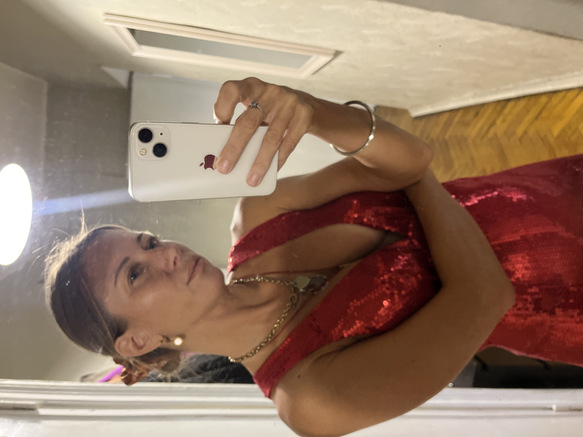 A woman is taking a selfie in a mirror while wearing a red sequined dress. She has her hair up and is wearing earrings, a necklace, a ring, and a bracelet. She is holding a white iPhone in her right hand. The background appears to be an interior space with a door and a mirror. There is no indication of the location being Moscow, Russia.