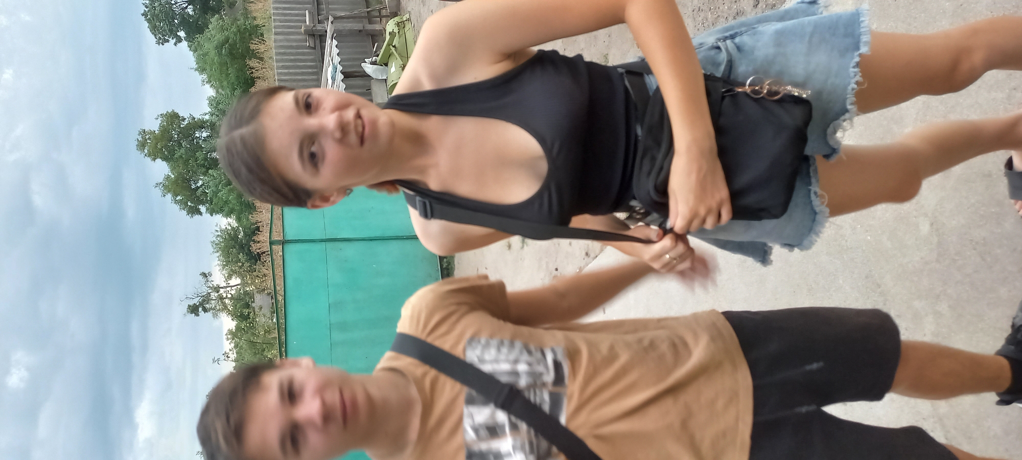 A young man and woman are standing outdoors in front of a teal-colored fence and some trees. The man, on the left, is wearing a tan t-shirt with a graphic print and dark shorts. He has a black messenger bag slung across his body. The woman, on the right, is wearing a black halter-top and a light blue denim skirt. She has a black crossbody bag around her waist. The woman is looking towards the camera and smiling slightly, while the man is looking in front of him. The background suggests a rural or suburban setting with a corrugated metal fence and some agricultural equipment visible to the right. The sky is overcast with clouds. This appears to be a candid outdoor photograph taken in Klyntsi, Ukraine.