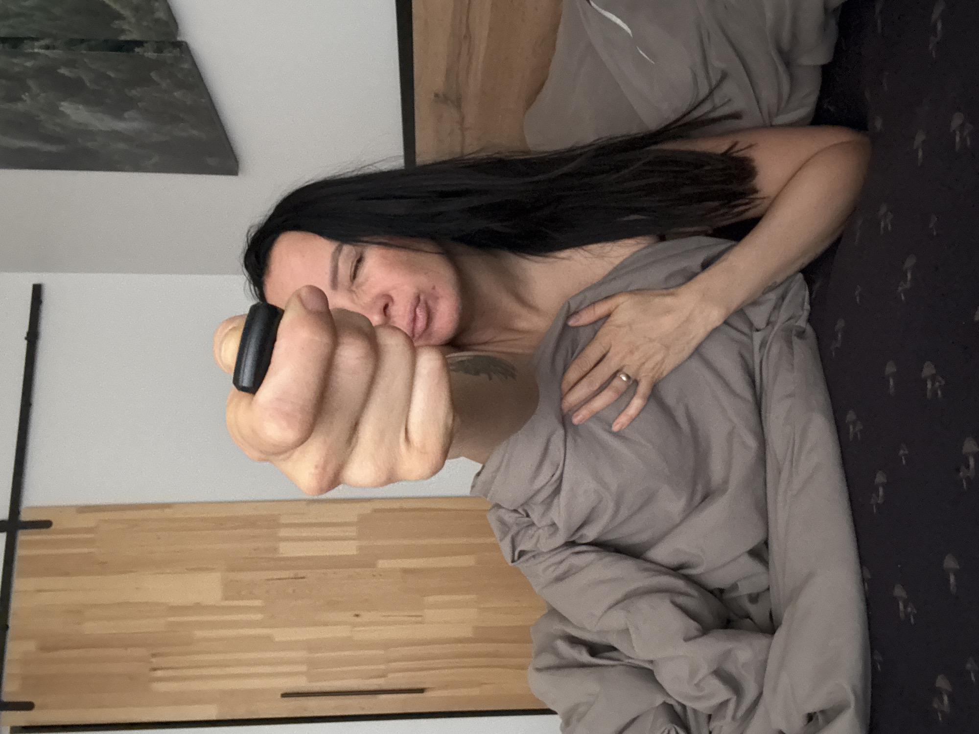 A woman with long, black hair is lying in bed, covered in gray bedding. She is holding a small, black object in her outstretched hand. A tattoo is visible on her shoulder. The headboard of the bed is wooden. A picture hangs on the wall above the headboard. To the left, there is a light-colored wooden surface with black metal hardware. A mushroom-patterned bed sheet is visible on the bed to the right of the woman. The location is Chaikovsky, Russia.