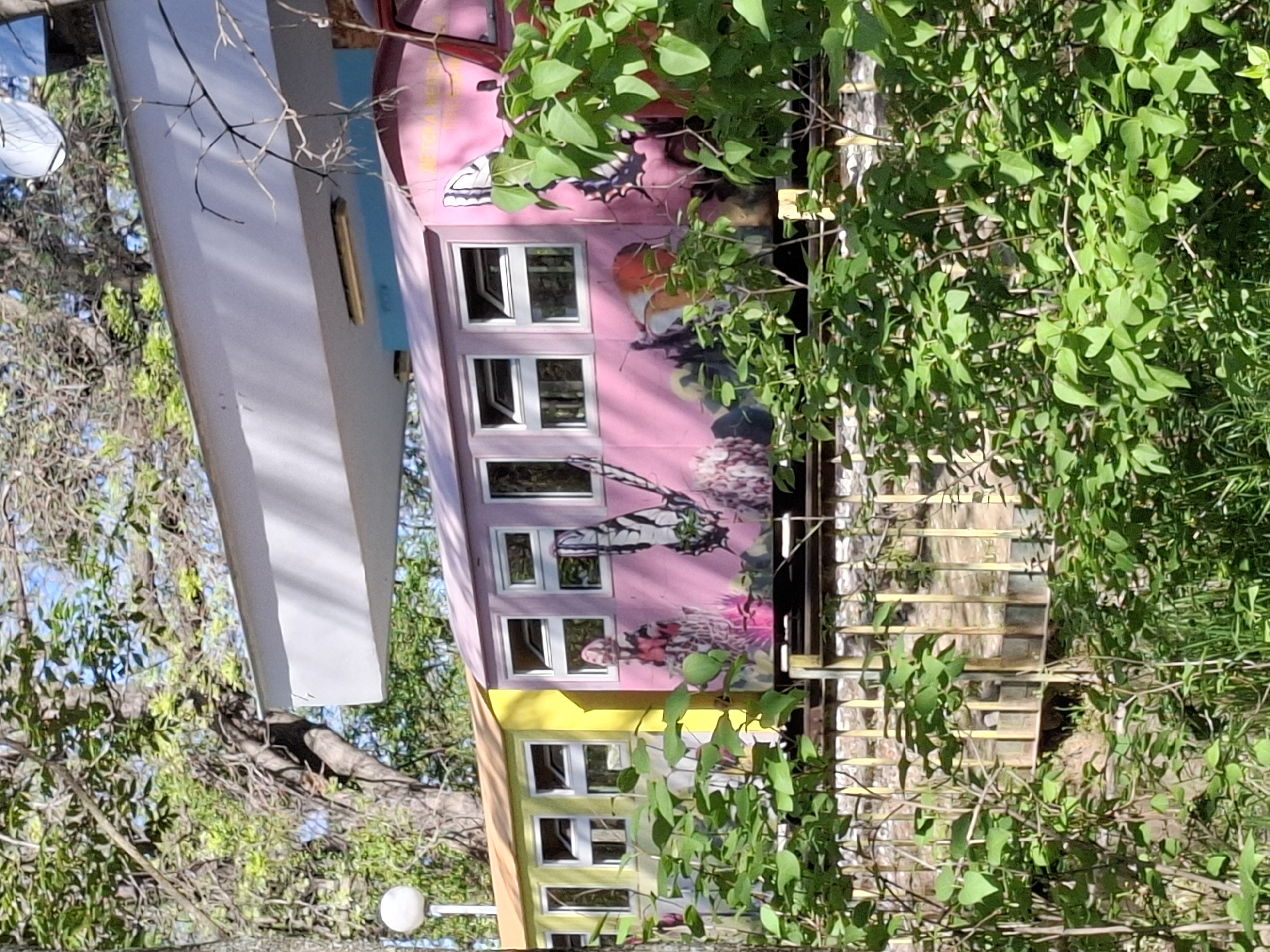 The image shows a colorful building with a pink and yellow exterior, adorned with paintings of butterflies and other nature-inspired designs. The building has multiple windows arranged vertically. A white, rectangular structure is attached to the side. Green foliage partially obscures the building and a wooden deck-like structure. The location is in a wooded area, likely in Plovdiv, Bulgaria, as the building is reminiscent of the "Art House" located on Nebet Tepe hill. No people are visible in the image. The scene depicts an artistic structure integrated into a natural setting.