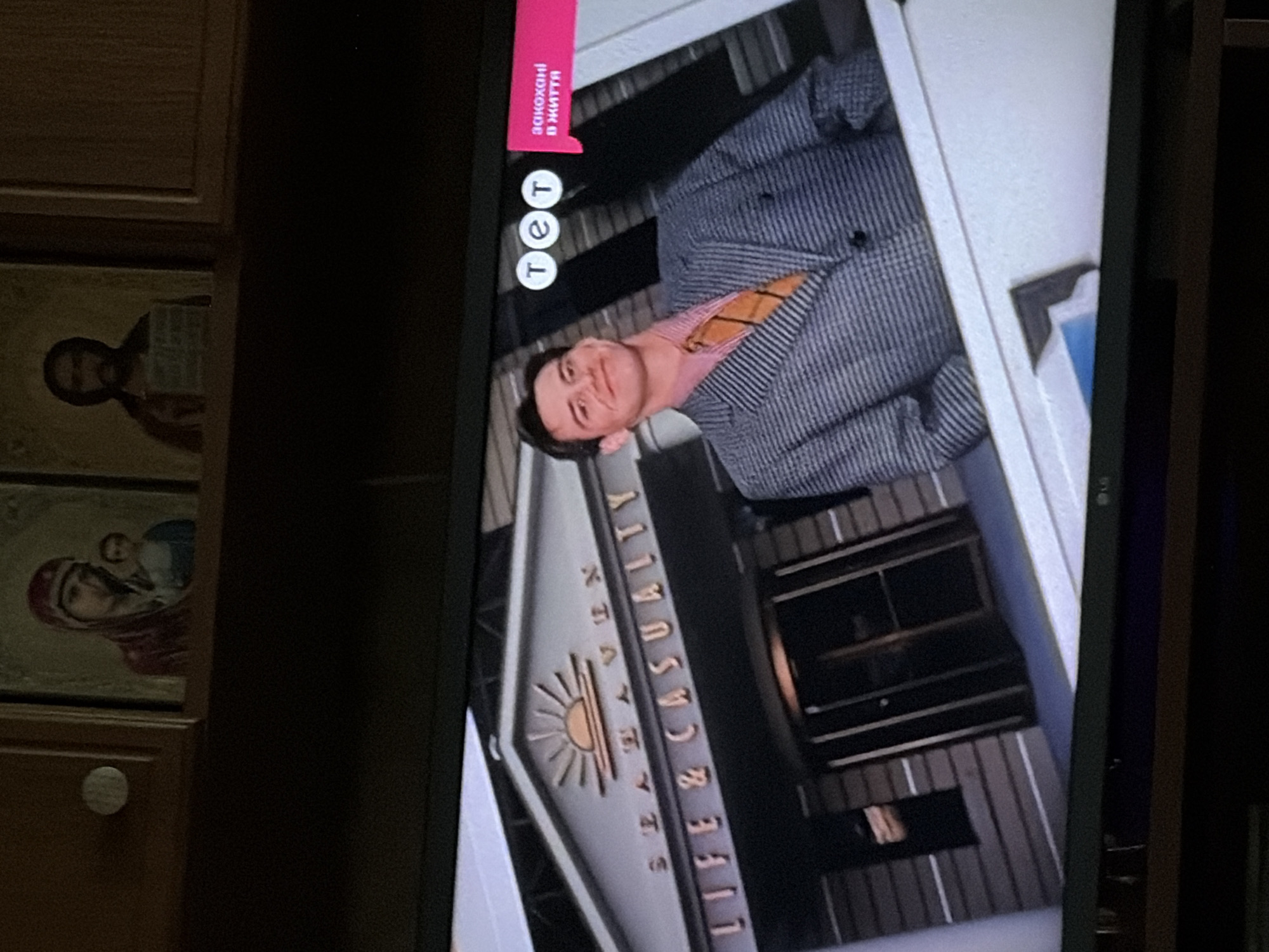 The image shows a television screen displaying a scene from a TV show or movie. In the foreground, a man wearing a grey plaid suit and an orange tie is looking at the camera with a slight smile. Behind him, a building facade is visible with the words "SEAHAVEN LIFE & CASUALTY" in gold lettering above an entrance. The building has a triangular pediment with a stylized sun logo. In the top left and right corners of the television screen, there are religious icons depicting figures in traditional attire. A logo with the letters "TET" appears in a circle, and a pink graphic with Ukrainian text is also visible. The setting appears to be an urban environment, possibly a hospital or office building.