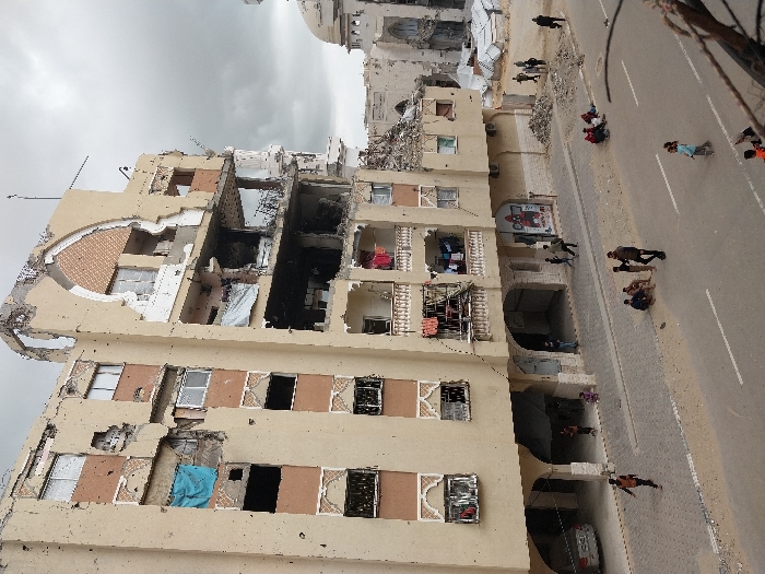 An elevated view captures a street scene in Khan Yunis, Palestinian Territory, under an overcast sky. The foreground is dominated by a severely damaged multi-story residential or commercial building, tan in color with reddish-brown decorative accents around windows and arches. This building exhibits extensive structural damage, particularly on its upper floors and right side, with large sections of its facade missing, exposing interiors, twisted rebar, and debris. Multiple windows are broken or entirely absent, and bullet or shrapnel impacts are visible across its surface. Blue tarpaulins are seen covering some openings, and laundry hangs from balconies on the lower intact floors.
To the right, in the background, stands another heavily damaged structure, characterized by a visible dome and pointed arches, consistent with a mosque, which appears partially collapsed. Piles of rubble are visible near its base.
The street below consists of asphalt with white lane markings. The adjacent sidewalk is paved, with areas of dirt and small piles of debris. Approximately twenty individuals are present on the street and sidewalk. Several small groups of men and boys are seen sitting or standing on the street and sidewalk. Individual children are observed walking and running on the street. Under the arched ground floor of the main building, a white sedan is parked, and more individuals are visible in the shadowed areas. The overall environment conveys widespread urban destruction and ongoing human presence amidst the damage.