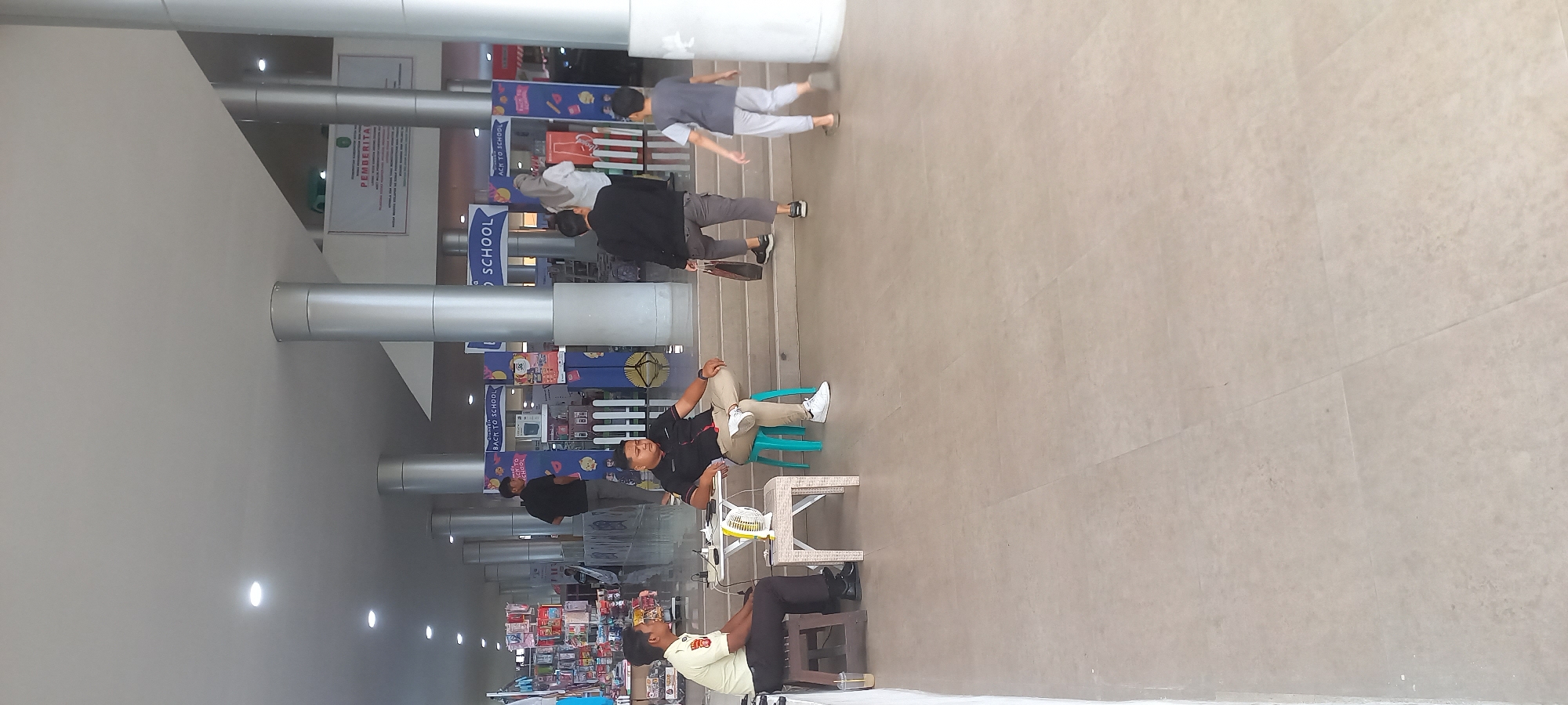This image depicts a wide-angle view of an indoor public space, likely a commercial building or a plaza, with elements suggesting a retail or event setting. The scene is dominated by large, cylindrical support columns and a light-colored tiled floor.
In the foreground, on the left side of the image, a man in a light-colored uniform with a badge is seated on a dark wooden stool. He is looking towards the left and appears to be interacting with a drone and electronic equipment on a white wicker table in front of him. To his right, another man, dressed in a dark collared shirt and light-colored pants, is seated on a teal stool. He is leaning back with his legs crossed and is looking towards the right. Both men appear to be stationary.
In the background, there are several other individuals. A man in a black t-shirt and gray pants is walking up a set of stairs. Further back, a man in a black jacket and gray pants is also ascending the stairs, carrying a brown paper bag. A woman in a light-colored hijab and white top is walking away from the camera, heading towards a display area. On the right side of the image, a young boy in a gray t-shirt and light gray pants is walking towards the right, away from the stairs.
The background displays various promotional booths or displays, with signage including "BACK TO SCHOOL" in multiple instances. There are also shelves stocked with goods, indicating a retail environment. A banner with Indonesian text, including the word "PEMBERITA," is visible further in the background, suggesting official information or announcements. The lighting is a mix of overhead artificial lights and ambient light filtering in from an unseen source. The overall impression is a busy, public area with ongoing activity. The presence of the drone and promotional displays suggests this could be an event, a product demonstration, or a retail area with ongoing sales promotions.
The description does not contain information that specifically places the scene in the city of Melayu, Indonesia.