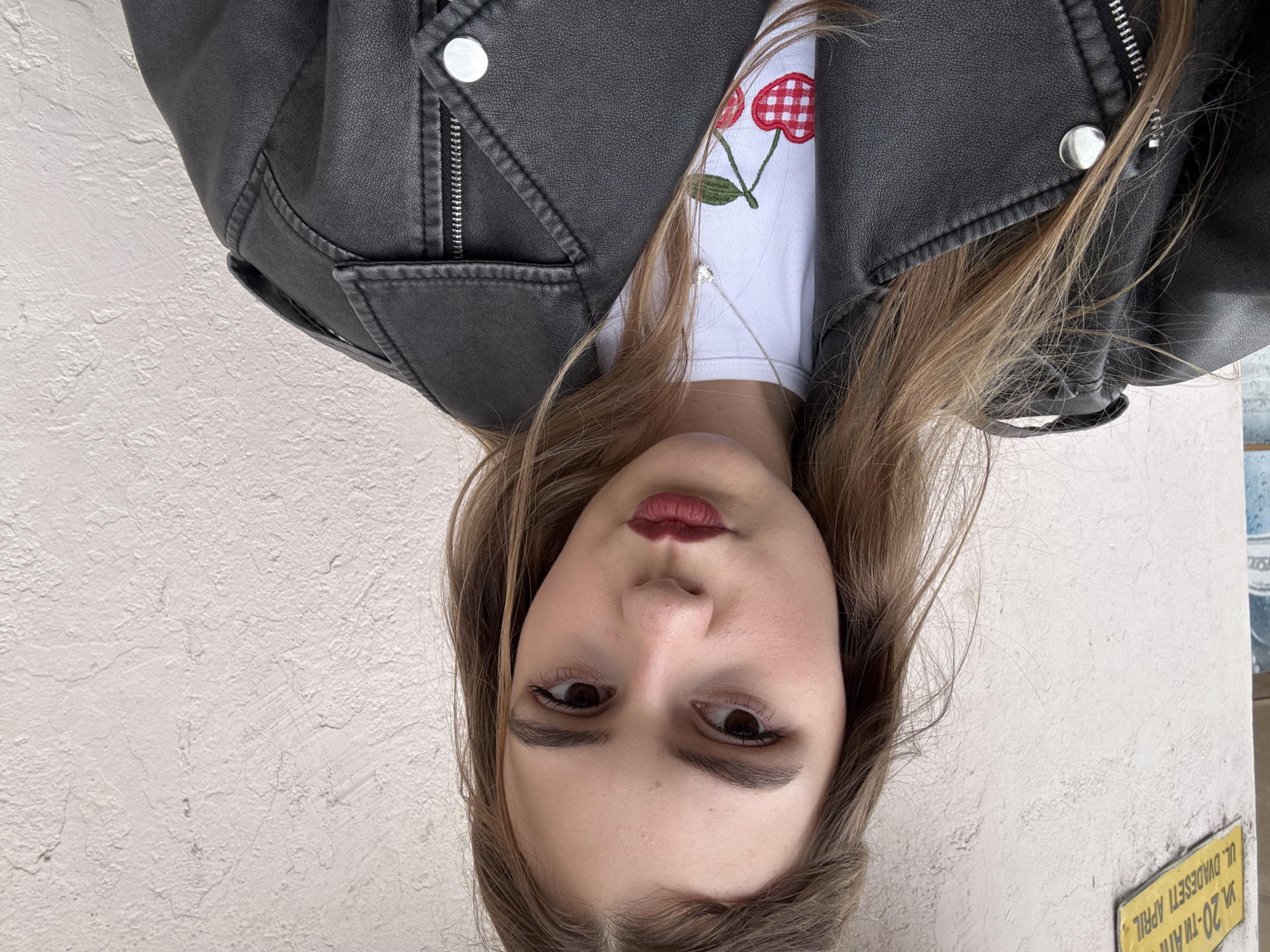 A young woman is centered in the frame, wearing a black leather jacket over a white t-shirt with a red cherry design. Her brown hair is parted and falls to either side of her face. She looks directly at the camera with dark eyes and a neutral expression, wearing dark lipstick. The background is a textured beige wall. A yellow sign is visible in the lower right corner, partially obscured, with text in Cyrillic indicating "ul. dvadesetti april" or "20th of April Street" which is an identifiable location in Panagyurishte, Bulgaria.