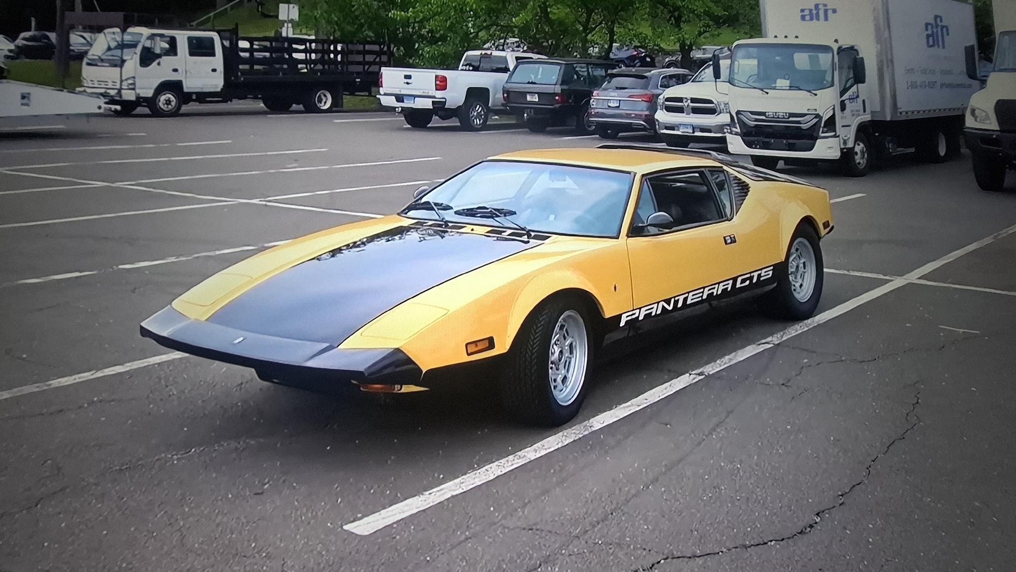 The image displays a bright yellow and black De Tomaso Pantera GTS sports car prominently parked in an asphalt parking lot. The car features a matte black hood, front spoiler, and a lower side stripe on which "PANTERA GTS" is written in white lettering. It is equipped with silver multi-spoke wheels and has a distinctive low, angular front design.
In the background, several other vehicles are parked. To the left, a white flatbed truck with a wooden-sided bed is visible. Further back, a white Chevrolet Silverado pickup truck is parked, alongside a dark SUV (possibly a Range Rover) and a grey SUV. To the right of the Pantera, a white Ram pickup truck and a white Isuzu box truck are visible. The box truck has "afr" and "Isuzu" branding on its side.
The scene takes place outdoors in a parking lot, indicated by the white painted parking lines on the asphalt pavement, which shows some cracks. Green trees and foliage are seen in the distant background. The lighting suggests it is daytime, possibly overcast, as there are no harsh shadows. All vehicles appear to be stationary, and no human activity or specific event is discernible.