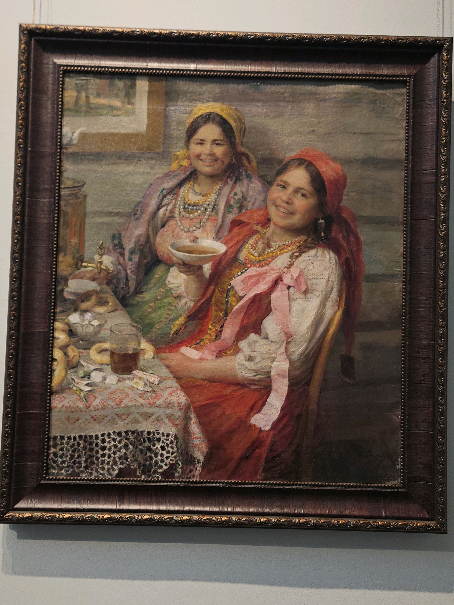 The image is a framed oil painting depicting two young women in traditional attire, seated at a table laden with food and drink. The woman in the foreground is wearing a red headscarf, a white blouse with lace trim, and a red dress, adorned with necklaces and a pink ribbon. She is holding a small bowl and smiling. The woman behind her wears a yellow headscarf and a colorful embroidered shawl over her dress. She also appears to be smiling. On the table, there is a samovar, a glass of dark liquid, a bowl of sugar cubes, and several round pastries, possibly bagels or kalachi. The table is covered with a patterned cloth and a lace runner. The background suggests an indoor setting with a window or doorway. The painting is framed in a dark, ornate wooden frame with gold accents.
This painting is located in the Ulyanovsk Regional Art Museum in Ulyanovsk, Russia.