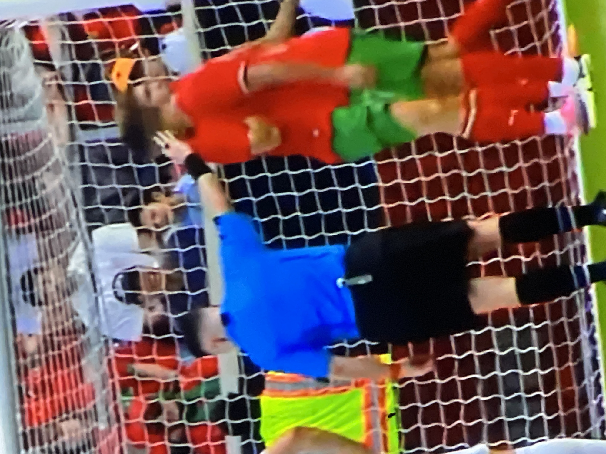 This image depicts a soccer match unfolding within a stadium in Casablanca, Morocco, focusing on the immediate vicinity of a goal. A male referee, clad in a bright blue short-sleeved shirt, black shorts, and black socks, stands center-left with his back partially to the viewer. A metallic clip or whistle is attached to his right hip, and a red band is visible on his right wrist. He extends his left arm, with his hand resting on the upper back or shoulder of a male player positioned to his right. This player wears a red short-sleeved jersey, green shorts, white socks, and light-colored athletic shoes; he has brown, wavy hair. Another player, similarly dressed in red and green, is partially visible to the right of the first player. A segment of a third individual, distinguished by a neon yellow-green bib, is visible on the far left. The prominent white mesh of a soccer goal net spans the middle ground, separating the field from the blurred background. Beyond the net, numerous spectators are discernible in the stands, exhibiting varied attire. The playing surface beneath the individuals is a vibrant green turf.