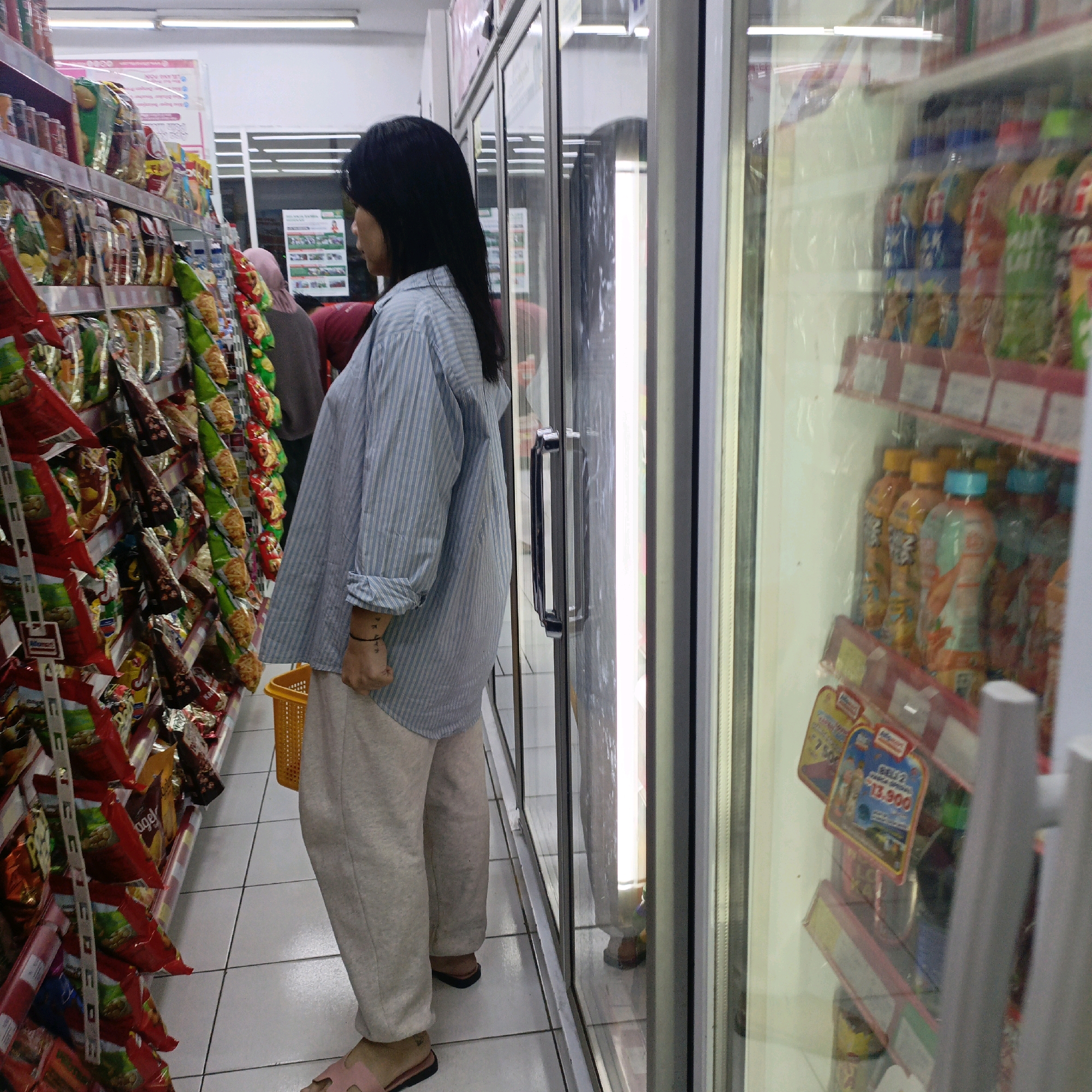 The image depicts an indoor scene inside a minimarket, possibly a branch of Indomaret or Alfamart, located in Salatiga, Indonesia. A young woman, facing away from the camera and towards the right, is browsing the shelves. She has long, dark hair and is wearing a light blue and white striped long-sleeved shirt and light-colored sweatpants. On her left wrist, there is a visible tattoo or bracelet. In her right hand, she is holding a yellow woven basket.
The minimarket environment includes aisles stocked with various products, predominantly snacks in colorful packaging. To the right of the woman, a refrigerated display case with glass doors is visible, containing rows of bottled beverages. The floor is tiled with a light-colored, square pattern. Reflections on the glass doors show other parts of the store, including shelves and possibly other customers. A prominent silver handle is attached to the glass door of the refrigerated display.
The overall lighting suggests a typical retail environment. The presence of a minimarket in Salatiga, Indonesia, is inferred from common signage and product displays seen in such establishments in the region.