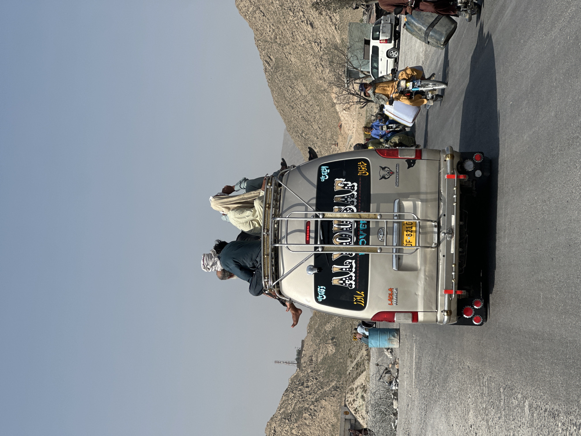 The image depicts an overloaded silver van, likely a passenger transport vehicle, with several people riding on its roof rack. It is in Quetta City Tehsil, Pakistan. The van is adorned with stickers and writing in Urdu and English. The license plate is visible, reading "DF 8210". Behind the van, other vehicles and people are visible on the road. A dry, mountainous landscape forms the backdrop, with a clear blue sky above.