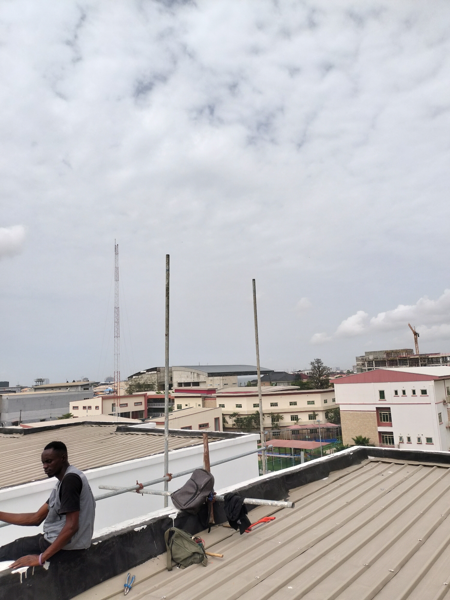 The image depicts a man sitting on the edge of a roof, likely in Moba, Nigeria. He is wearing a dark t-shirt and a light gray vest. Scaffolding is set up nearby, along with a gray backpack, a green satchel, and tools including pliers and a small hammer. The roof is corrugated metal with black flashing. In the background are several buildings, including one with a red roof and white walls. Further in the distance, a tall communication tower and a construction crane can be seen against a cloudy sky.