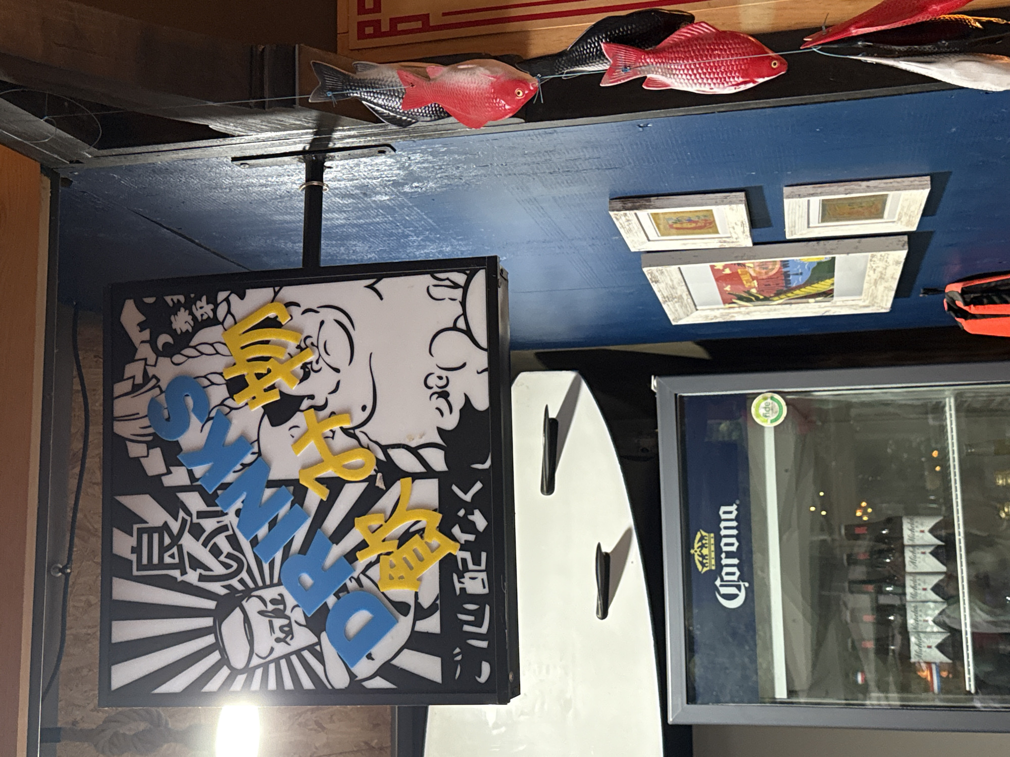 The scene depicts an interior section of an establishment in Puebla, Mexico, characterized by a dark blue wall and eclectic decorations. A square light-box sign, framed in black, occupies the upper left, featuring a black-and-white graphic with a Japanese aesthetic, incorporating blue letters spelling "DRINKS" and yellow Japanese characters reading "飲物" (drinks) and "ご心配なく" (don't worry). A stylized sake cup labeled "sake" and a cartoon pig figure are part of the design. Below the sign, a commercial glass-door refrigerator with a dark gray frame displays the "Corona" brand logo at the top and contains multiple bottles of "Michelob" beer. A green circular "fide" sticker is visible on the refrigerator door. To the right, the dark blue wall features a vertical string of four decorative fish, alternating between red and a darker/silver color. Below the fish, three framed pictures with distressed white frames are mounted on the blue wall, depicting colorful, abstract imagery. An orange object, possibly a safety vest, is partially visible hanging in the lower right portion of the blue wall. Wooden structural elements are present in the upper section of the image. No individuals are visible.
