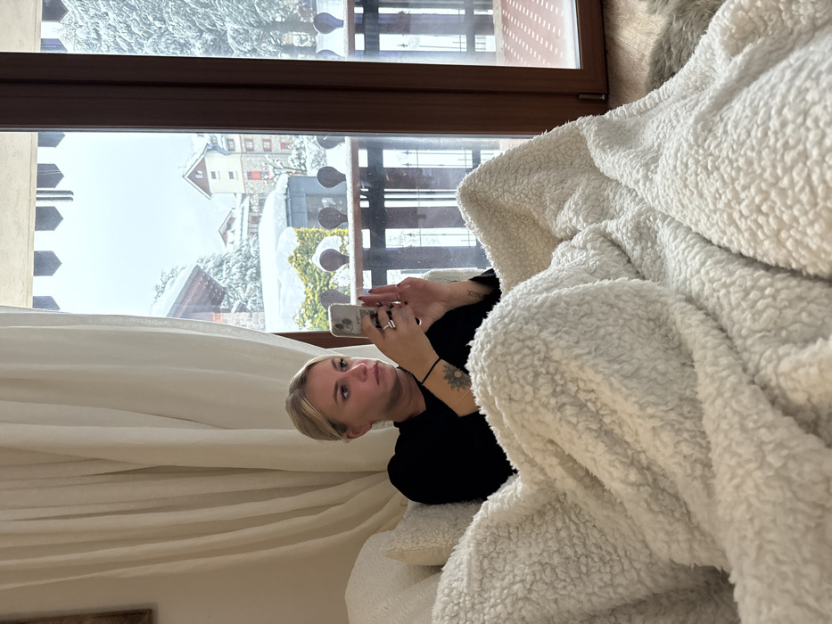 A fair-skinned woman with blonde hair holds a smartphone, possibly taking a picture. She is indoors, near a window displaying a snowy landscape and buildings. Part of her body is covered by a white, fluffy blanket or throw. She wears a black long-sleeved shirt and has a tattoo on her left arm. There is a curtain visible in the background. The photograph has been rotated slightly.
