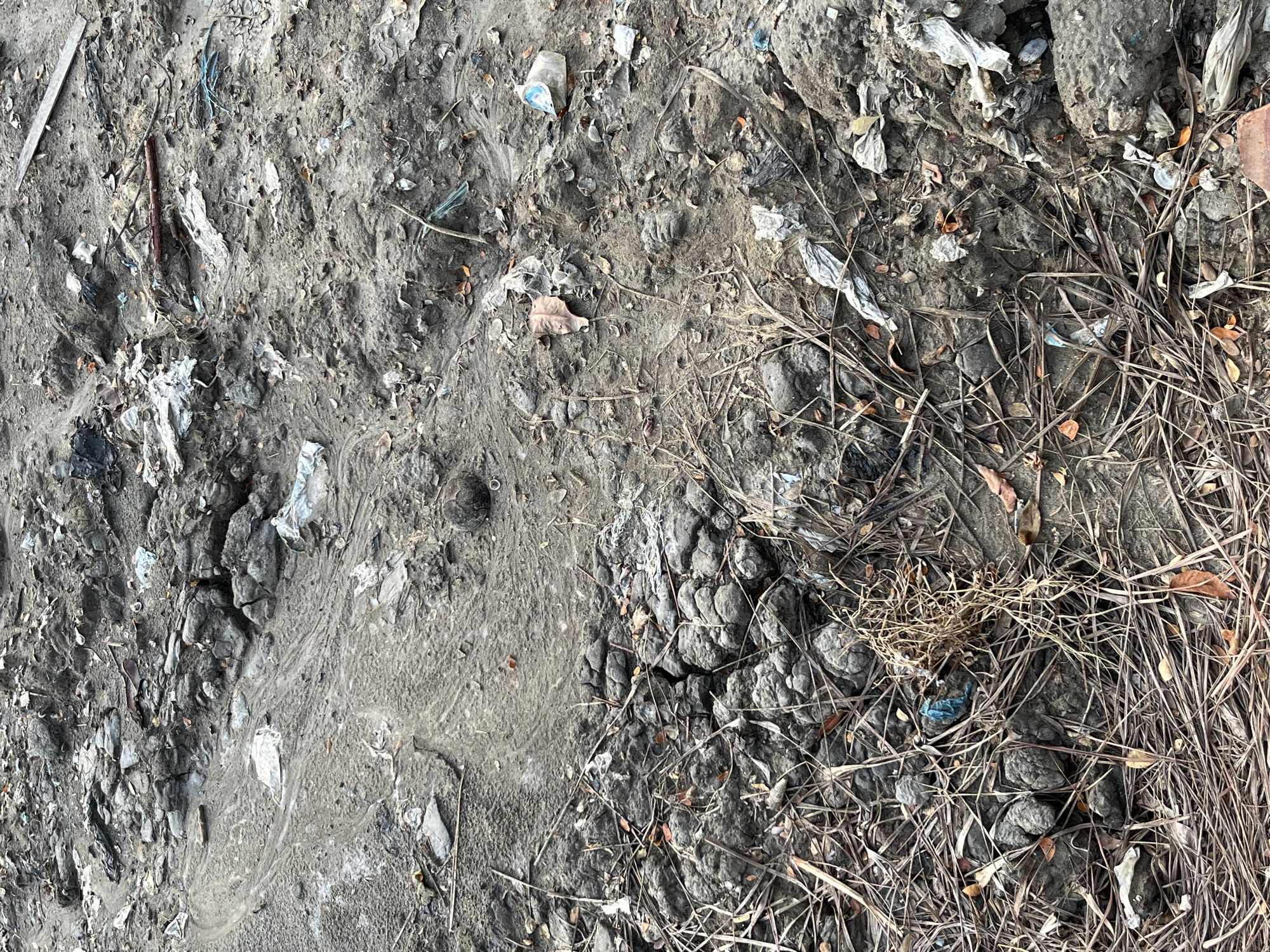 The image depicts a close-up, overhead view of a muddy, debris-strewn ground surface. The ground is uneven, with clumps of dark, cracked mud and loose soil. Scattered across the surface are various types of refuse, including plastic wrappers, bits of fabric, dried leaves, and small twigs. A few larger, darker, rounded objects, possibly stones or compacted mud clumps, are visible, particularly in the lower left portion of the image. The overall impression is one of neglect and accumulation of waste in an outdoor, possibly undeveloped or unmaintained area. The scene is devoid of any human presence or discernible action. The context provided indicates this scene is located in Lhokseumawe, Indonesia.