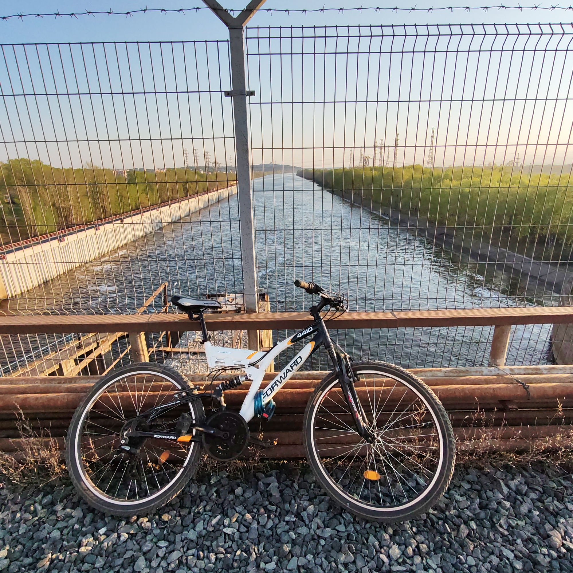 A bicycle is parked on a bridge over a canal in Chaikovsky, Russia. The bike is a black and white "Forward 4410" mountain bike. The bridge has a metal railing and a fence topped with barbed wire. A concrete canal runs below the bridge, with vegetation on both sides of the canal. Electrical transmission towers are visible in the distance. The ground around the bridge is covered in gravel. The weather appears to be clear and sunny.