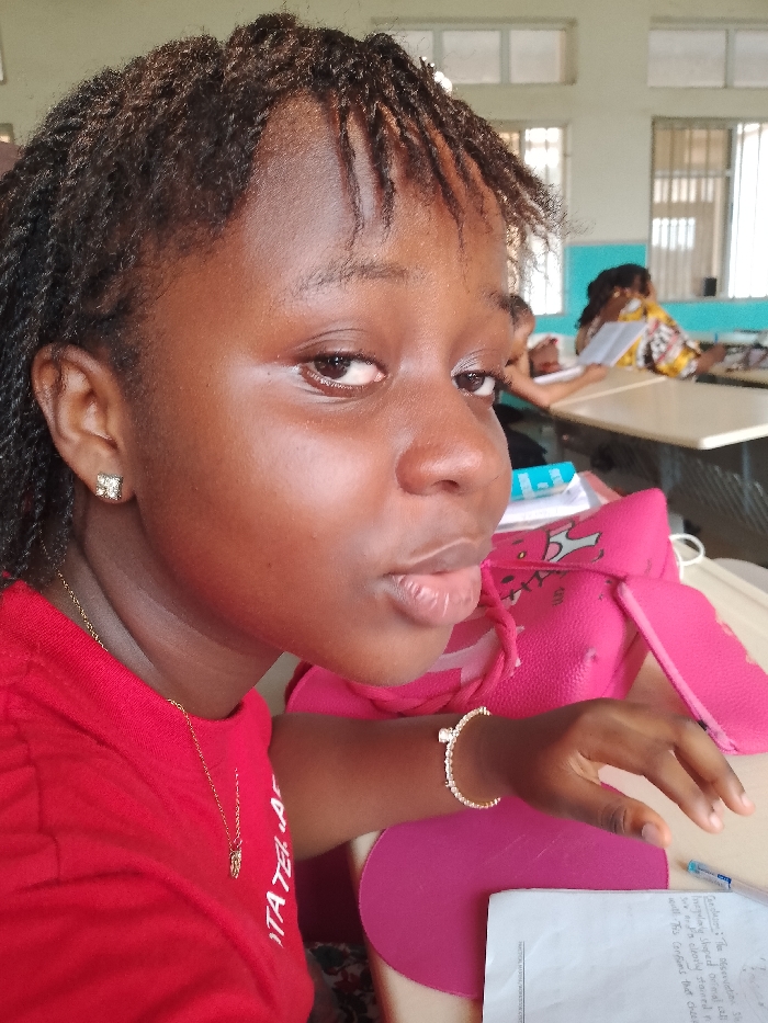 The image shows a young woman in a red shirt, wearing gold jewelry (earrings, necklace, bracelet) sitting at a desk. She is in what appears to be a classroom setting, with other students visible in the background. She is looking at the camera. There is a pink bag next to her, as well as school-related items like paper and a pen. The setting is in Ogboko, Nigeria.