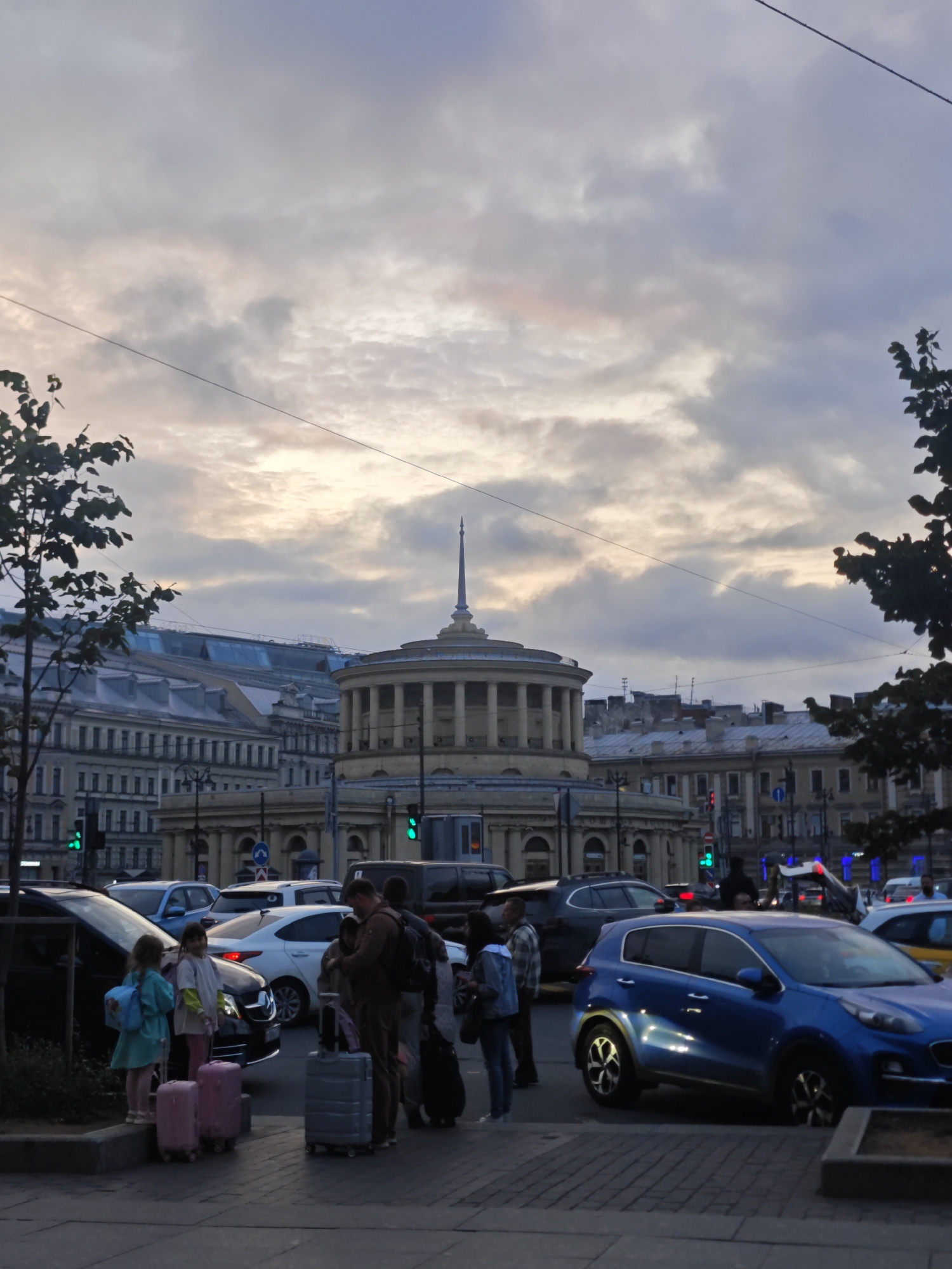 This media file captures an urban street scene in Saint Petersburg, Russia, at dusk. The sky is overcast with dramatic clouds displaying hints of pink and orange from the setting sun, suggesting a cool, possibly humid evening.
The foreground features several people with luggage, indicating travel or an imminent departure/arrival. Two young girls in light-colored clothing stand near a black car, each with a pink suitcase. Behind them, a group of adults are gathered, some with suitcases, suggesting they are waiting for transportation or meeting someone. One person, possibly a man, is dressed in darker clothing and appears to be looking at his phone. Another woman, in a light-colored jacket, stands beside him.
The midground is dominated by a traffic intersection with several cars, including a distinctive blue SUV in the right lane. Traffic lights are visible, with at least one showing green, suggesting traffic is moving. The architecture in the background is characteristic of Saint Petersburg, with grand, classical buildings. A prominent circular building with a dome and columns stands out, possibly a historical landmark. Other buildings feature mansard roofs, a common architectural feature in the city. Overhead, power lines crisscross the sky.
The overall mood is one of transient activity within a grand, historic urban environment. The low light conditions and the presence of travelers create a sense of a journey in progress. There is no visible text in the media file.