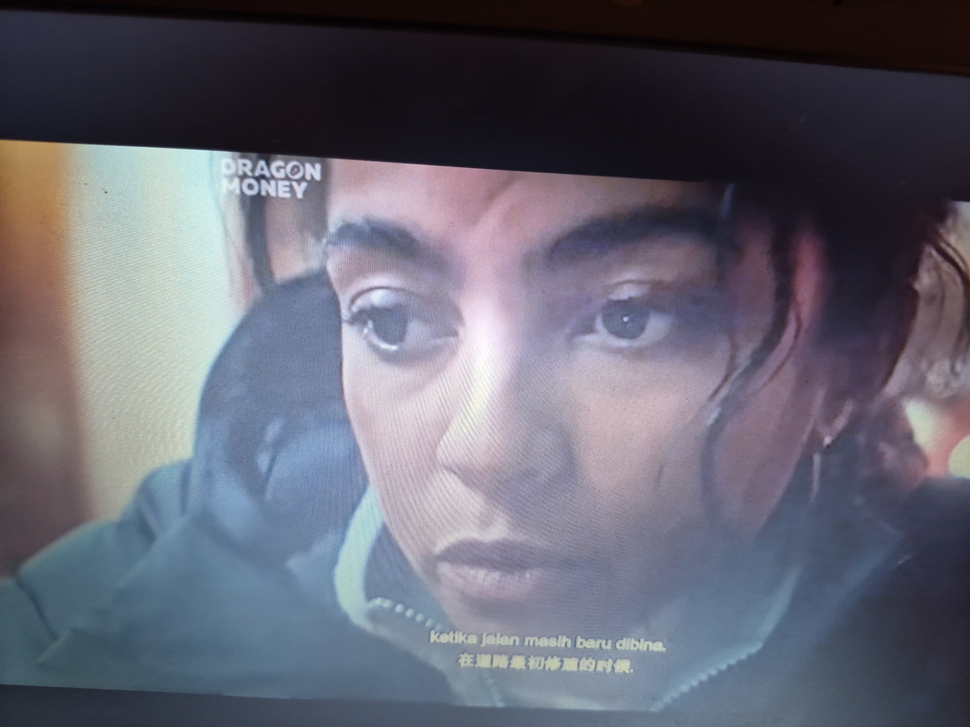 The image is a close-up, slightly blurry, medium-shot of a young woman's face, positioned centrally. She has dark, curly hair and is looking downwards and slightly to her left. Her expression appears contemplative or sad. The lighting is dim, with warm tones on her skin and cooler tones on her clothing. Text overlays are visible in the lower portion of the image, appearing to be subtitles. One line of text is in Roman script, "Ketika jalan masih baru dibina," and below it is text in Chinese characters. In the upper left corner, the words "DRAGON MONEY" are visible in white text. The background is out of focus, showing indistinct shapes and colors, suggesting an indoor environment, possibly a room or a vehicle. There is no discernible information that would place this image in the city of Sandwell, United Kingdom.
