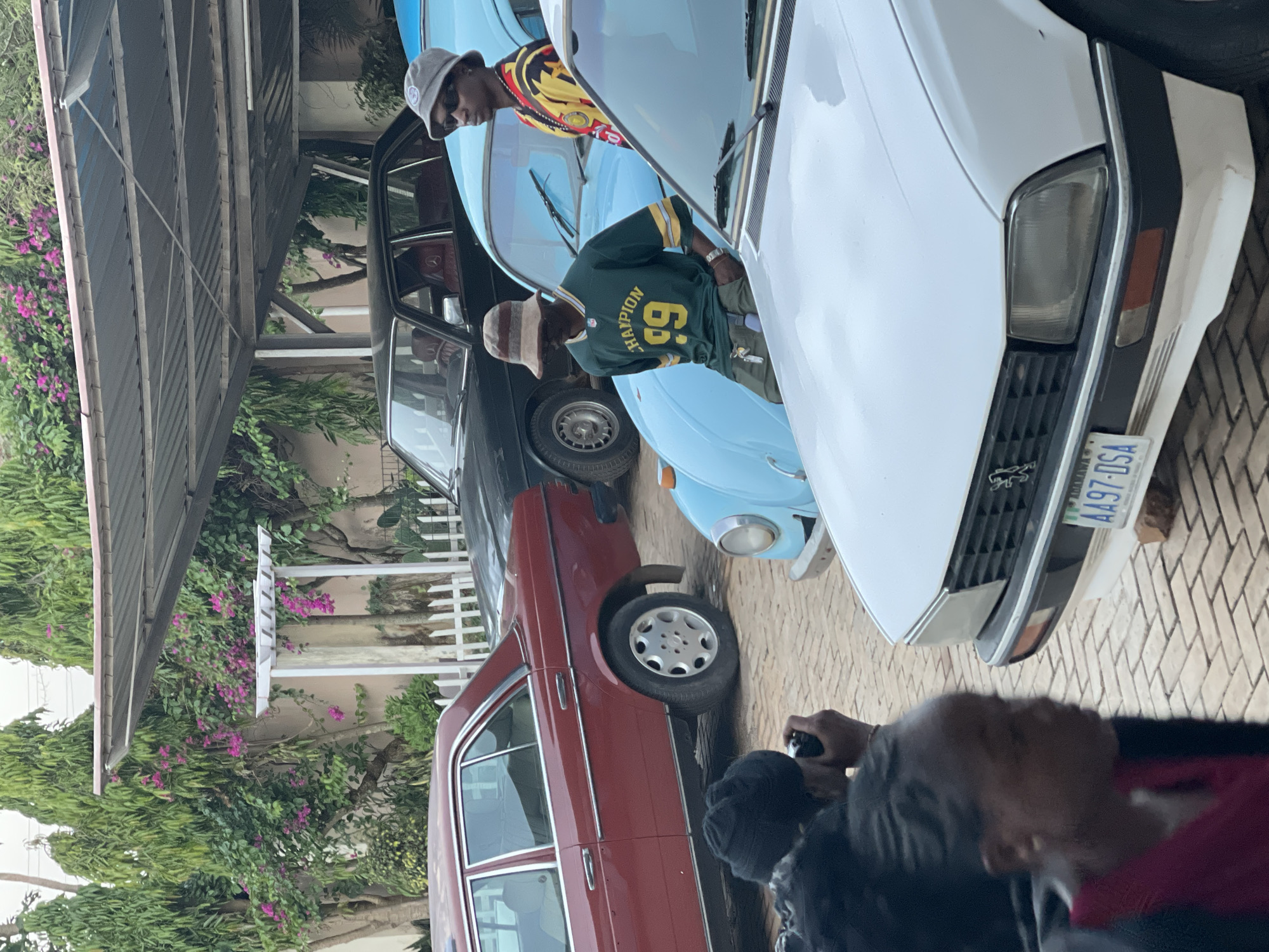 The image depicts an outdoor scene in Jos, Nigeria, featuring several individuals and vintage cars. Two men stand near a light blue Volkswagen Beetle, while a white Peugeot with a license plate reading "A497-DSA" is positioned in the foreground. A red Mercedes-Benz sedan is partially visible behind the Volkswagen. A dark-colored vehicle is parked to the left, partially obscured by the other cars. The environment includes a paved area, a white picket fence, and flowering vegetation, suggesting a residential or semi-urban setting. A person holds a camera, potentially documenting the scene. The scene appears to be centered around the vintage cars and the people interacting with them.