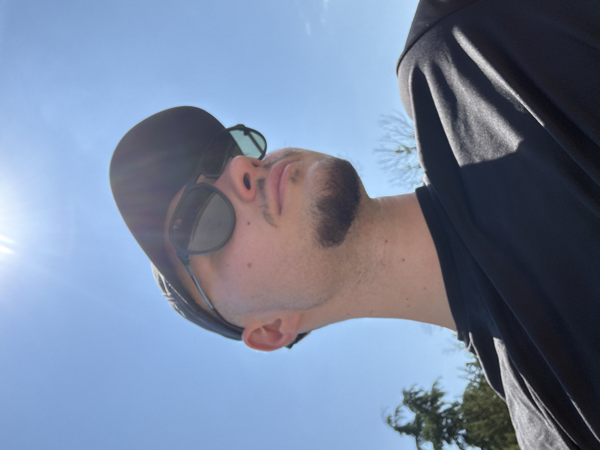 This is a low-angle, close-up shot of a person's face and upper body against a bright blue sky, suggesting it is daytime and the weather is clear and sunny. The person is wearing a black baseball cap and black sunglasses. They have short, dark hair and some facial hair. They appear to be looking upwards, possibly towards the sun, with their mouth slightly open. The lighting indicates it is likely midday, with the sun creating a strong glare on the left side of the image.
The person is wearing a black t-shirt or similar dark garment. The background is primarily the sky, with a hint of green foliage and tree branches visible in the upper right and lower right corners, suggesting an outdoor setting, possibly a park or garden. The image seems to be a selfie, given the perspective and the focus on the person's face. There is no visible text in the image. The overall mood is casual and relaxed, perhaps capturing a moment of enjoying the outdoors on a bright day. The location context of Sofia, Bulgaria, does not provide specific visual cues within the image itself, but suggests the general geographical setting.