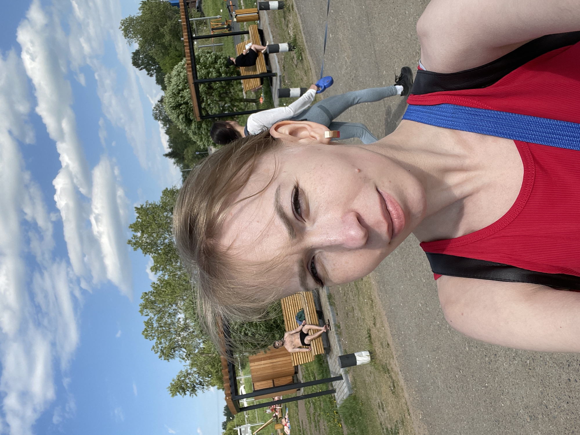A selfie was taken at a park in the city of Perm, Russia. The person taking the photo is wearing a red and black tank top with a blue strap. Two other people are visible in the background. One is wearing a white shirt and grey pants and appears to be exercising. The other is laying on a bench. The location appears to be a public park with exercise equipment. The weather is sunny with some clouds.