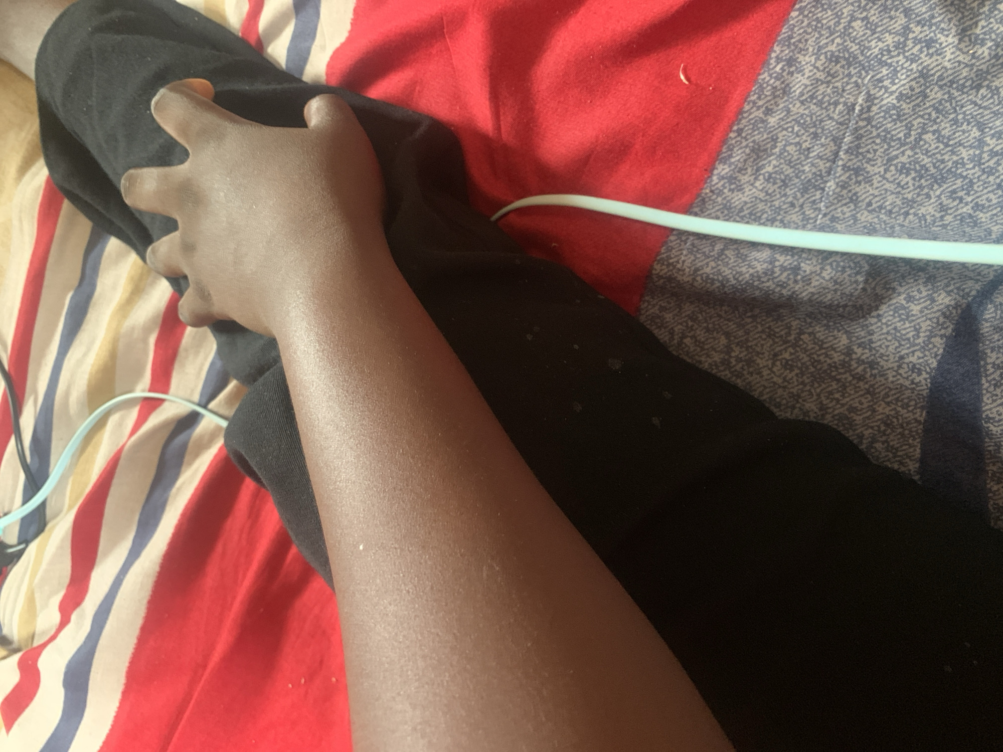 The image shows a close-up view of a person's arm and hand resting on black fabric. The arm is dark-skinned and appears to be slightly goosebumped. The hand is positioned with fingers slightly splayed, resting on the black fabric. The background consists of a striped blanket with red, white, and blue stripes. A thin, light blue cable is visible in the foreground, curving across the blanket and near the arm. The scene appears to be indoors, likely on a bed or sofa, in Jauro Yinu, Nigeria.