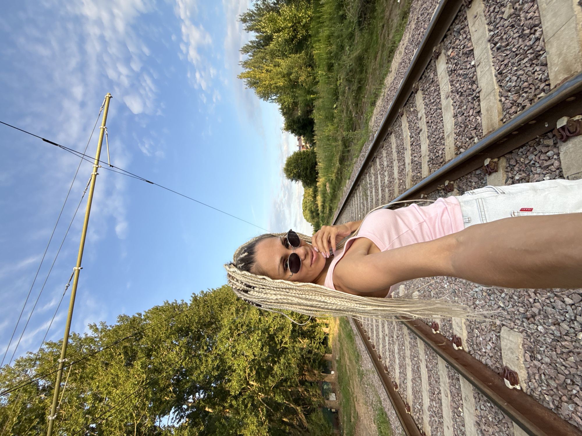 A woman with blonde braided hair and sunglasses stands on a railway track, holding a phone out to take a selfie. She is wearing a pink tank top and white shorts. The railway tracks stretch into the distance, flanked by trees on either side. The sky is blue with scattered clouds. The location is identified as Rusanovo, Russia.