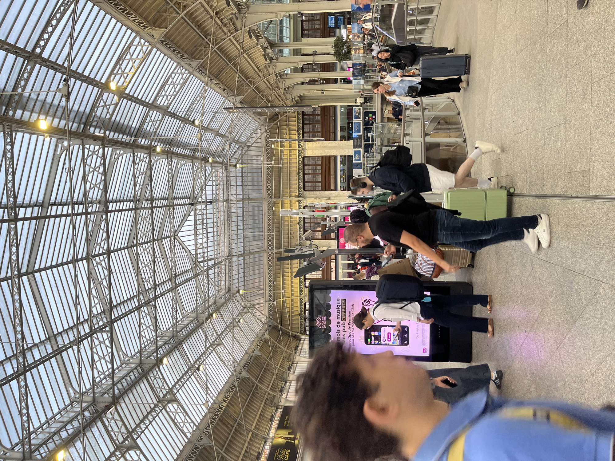 The image captures the interior of a large, busy train station with a high, arched glass and metal roof structure. The natural light filtering through the roof illuminates the space, with artificial lights also visible. People are seen walking in various directions, many carrying luggage, suggesting a travel context. Some individuals are blurred due to motion. In the foreground, a person in a blue shirt is partially visible, out of focus. A large digital advertisement for Spotify Premium is prominent in the mid-ground. People are dressed in casual attire. The architecture of the station, with its exposed beams and intricate metalwork, is a dominant feature. The overall scene depicts a moment of transit and activity within a grand public space, likely a major transportation hub. This scene is identifiable as Gare du Nord in Paris, France, due to its distinctive glass roof architecture and the visible advertisements and station signage characteristic of this Parisian landmark.