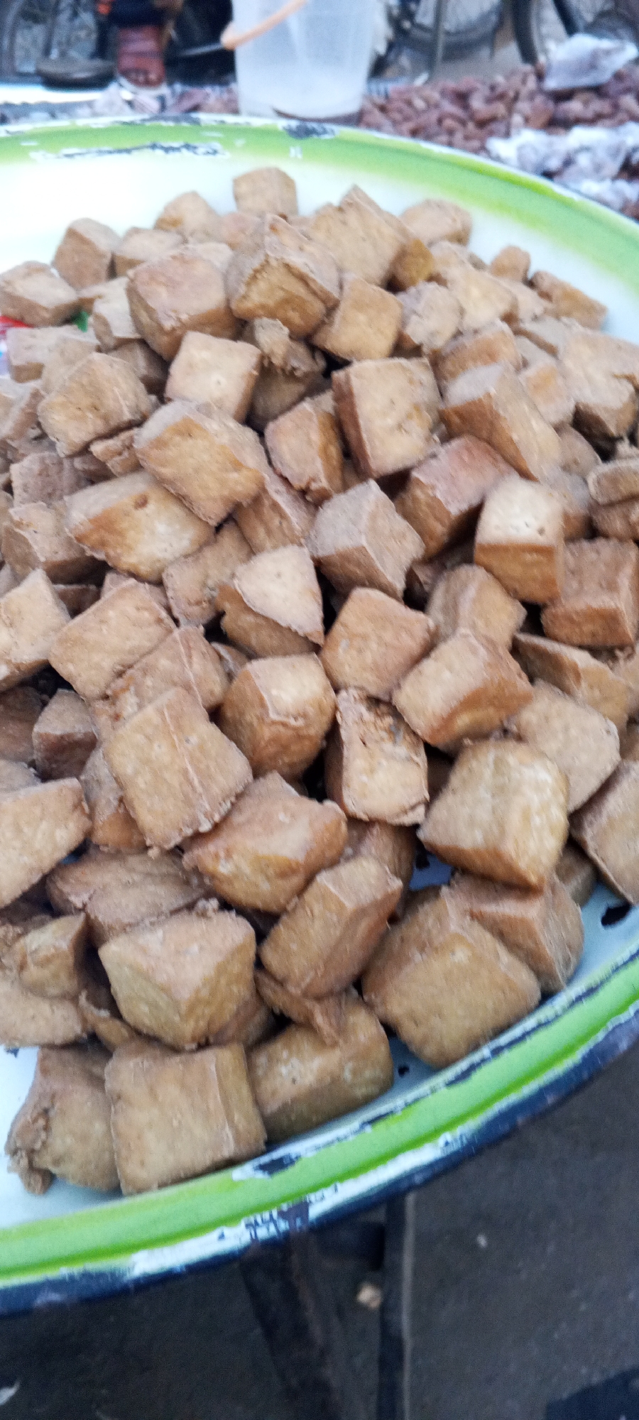 In Zango-ndaji, Nigeria, a metal tray, ringed with green, overflows with small, fried tofu cubes. The tofu is golden brown, packed densely, and dominates the foreground. In the indistinct background, hints of a bicycle, a white plastic bucket, and other objects suggest an outdoor market setting. No people are clearly visible. The focus is sharply on the mound of fried tofu, possibly for sale.