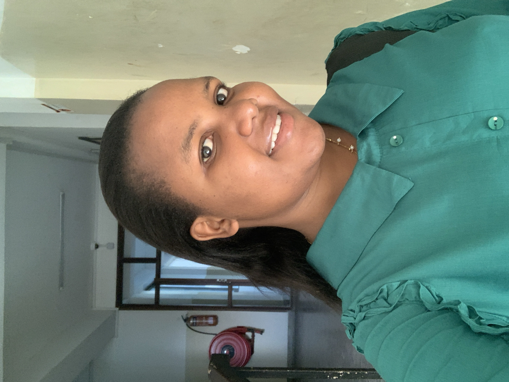 A woman is standing in a stairwell in Dar es-Salaam, Tanzania. The stairwell is dimly lit, with a red fire hose and extinguisher visible on one wall. The woman is wearing a green blouse and a pearl necklace. Her hair is pulled back, and she is smiling at the camera.