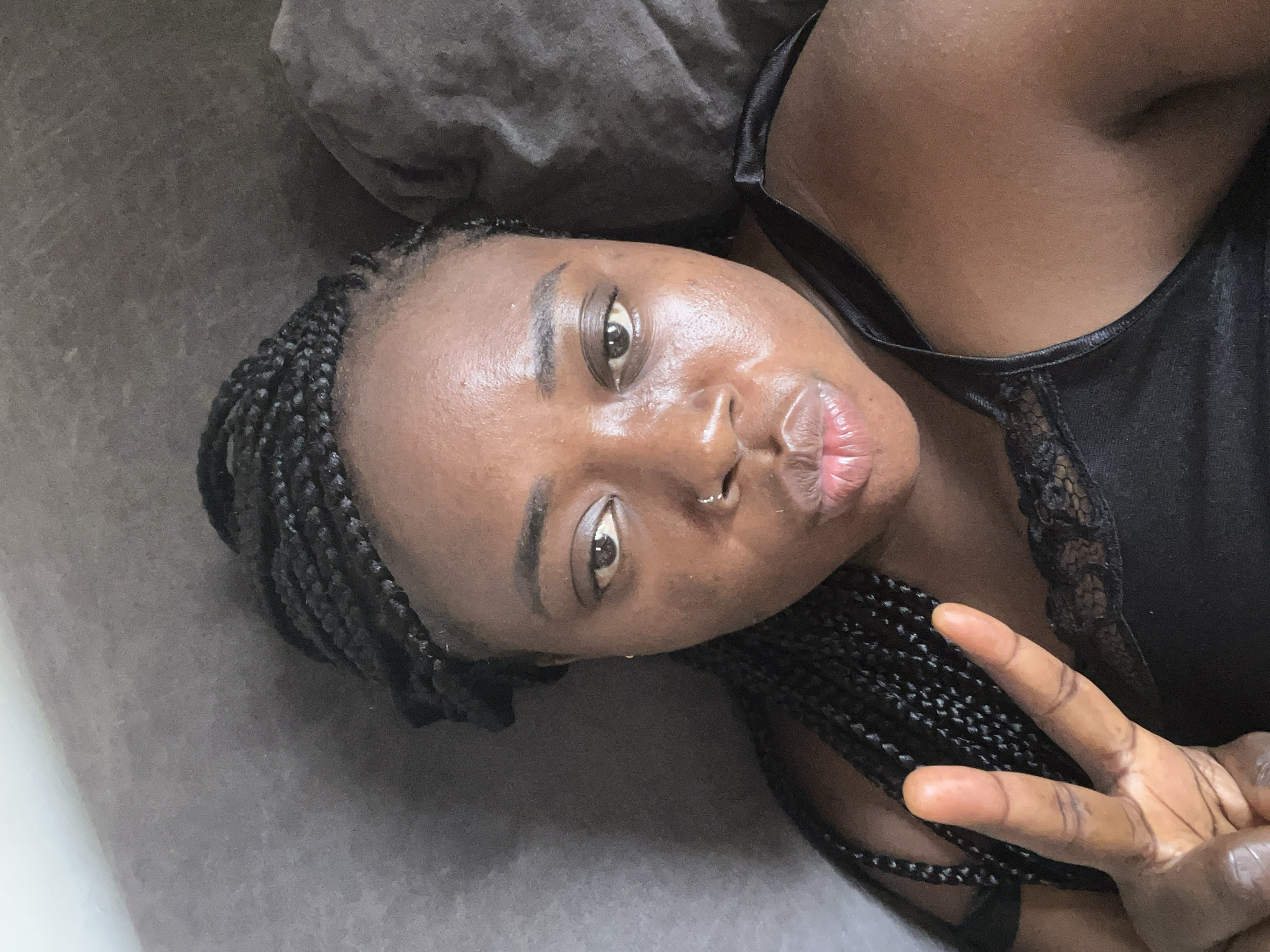 A close-up, eye-level shot depicts a dark-skinned woman with braided hair lying on a gray surface, possibly a bed or sofa, in an indoor setting. She is wearing a black top with lace details and is making a peace sign with her right hand near her face. Her facial expression includes pursed lips and a direct gaze at the camera. The background consists of a gray pillow and a glimpse of a white surface, possibly a wall. The image does not provide any visual clues indicating the location is Abuja, Nigeria, or any specifics about the environment beyond the immediate setting.