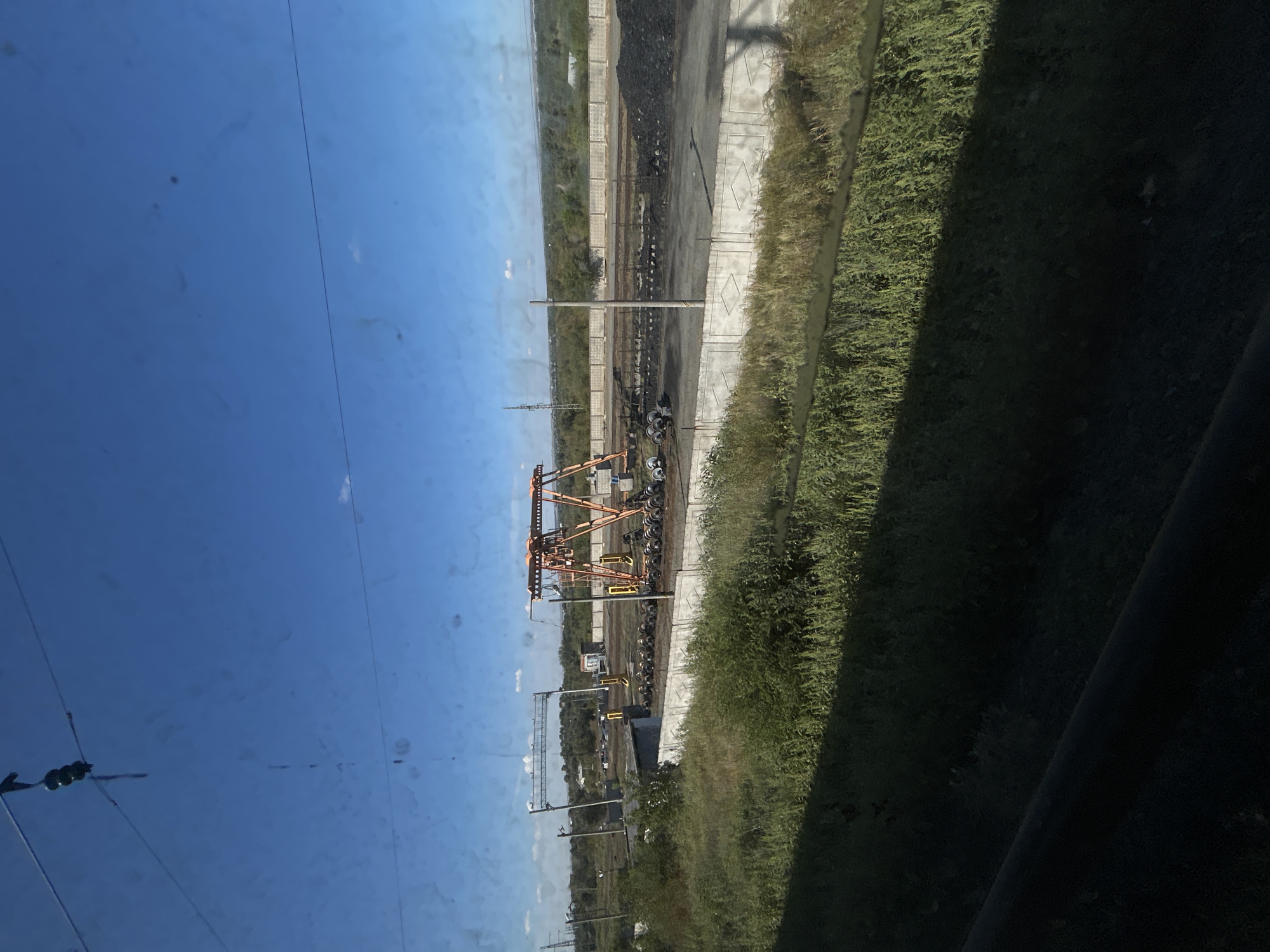 The image depicts an outdoor industrial scene under a clear blue sky with a few scattered clouds. In the foreground, tall green grass and vegetation are visible, casting a shadow across the lower portion of the frame. Beyond the vegetation, a concrete barrier runs horizontally across the middle ground, separating it from an industrial area.
Within the industrial area, there are railroad tracks and piles of dark material, possibly coal or ore. A prominent orange gantry crane stands in the center, with its boom extended. Several cylindrical objects, resembling rolls of material or large wheels, are stacked in rows near the crane. In the background, a concrete wall encloses more of the industrial site, and further in the distance, trees and a power pylon can be seen against the horizon. The overall impression is of a functional industrial facility.
Based on available information, this scene is consistent with the industrial areas within the city of Orsk, Russia, which is known for its mining and metallurgical industries.