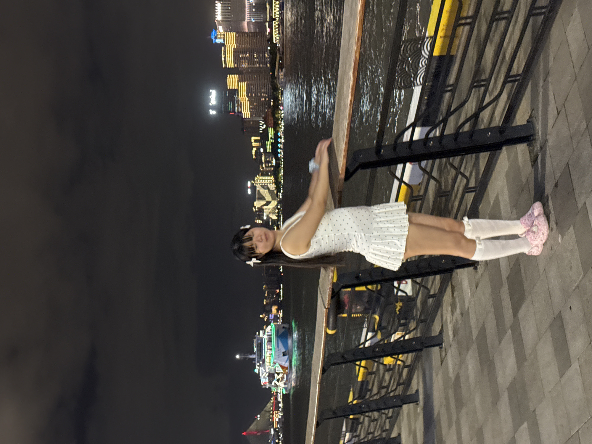 A young woman stands on a paved walkway at night, posing with her arms resting on a wooden railing. She wears a white polka-dot dress, white knee-high socks, and pink shoes. In the background, a brightly lit boat sails on a river, with the Shanghai skyline, featuring illuminated skyscrapers, visible across the water in Hongkou District, China. The sky is dark and cloudy.