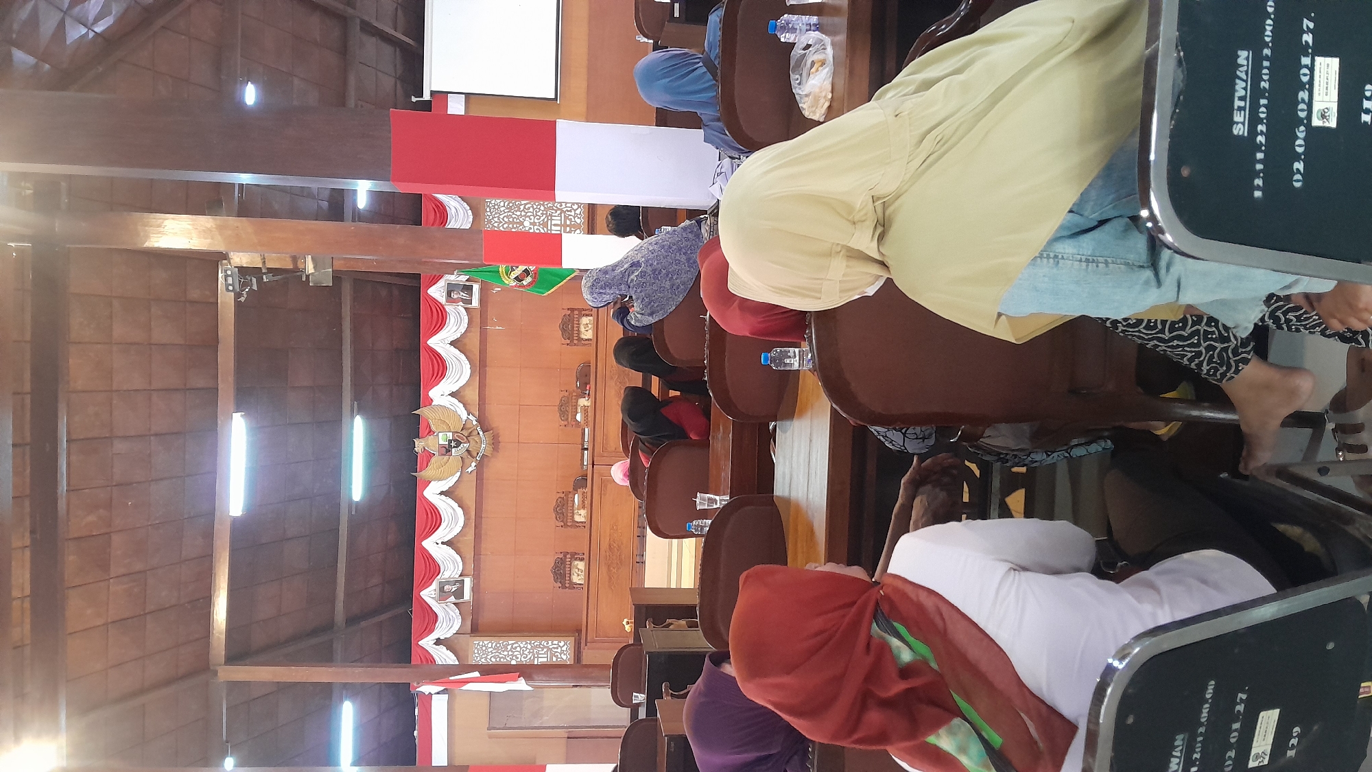 The image depicts an indoor scene with a wooden roof and wooden pillars, possibly a meeting room or hall. In the background, there is a wooden dais with chairs and a large wooden panel featuring an emblem and framed portraits. A flag with green and white stripes is also visible. The foreground and middle ground are occupied by rows of chairs where people, mostly women wearing headscarves, are seated. Some chairs have water bottles on them. The overall atmosphere suggests a formal gathering or presentation. Based on the context of the provided crops which show text such as "SETWAN" and dates in DD.MM.YYYY format which are common in Indonesian administrative documents, this scene is likely from an administrative building in Blora, Indonesia.