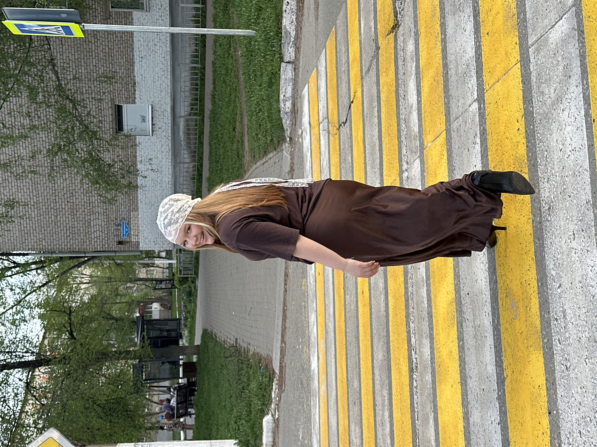 A woman wearing a long brown dress, a white lace cap, and black heels is walking on a yellow and white crosswalk in the city of Chaikovsky, Russia. The crosswalk is situated near a sidewalk, grassy area, and buildings. The woman is smiling at the camera. A traffic sign is visible in the top left corner of the image.