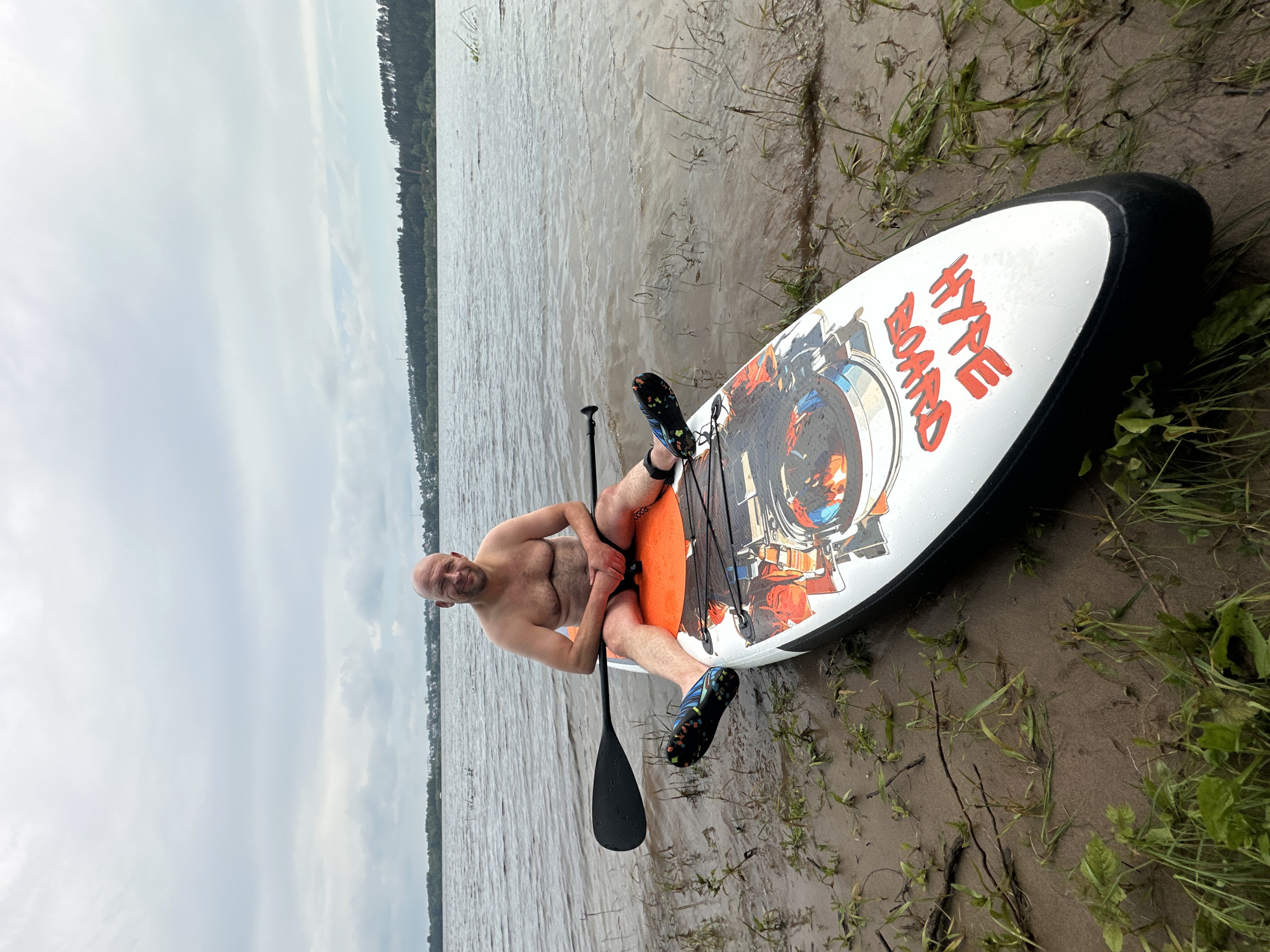 A bald, fair-skinned man with dark hair on his torso is sitting on an orange and white paddleboard partially submerged in shallow water near a muddy shoreline. He is wearing black swim trunks and black and blue water shoes. His hands are resting on his knees, and he is looking at the camera with a slight smile. A black paddle rests across the paddleboard behind him. The paddleboard has orange text that reads "HYPE BOARD" and graphics of a person DJing. The shoreline is covered in wet sand and green grass. In the background, there is a body of water with a distant treeline and buildings. The sky is overcast. The location is the Volga River in Noviy, Russia.