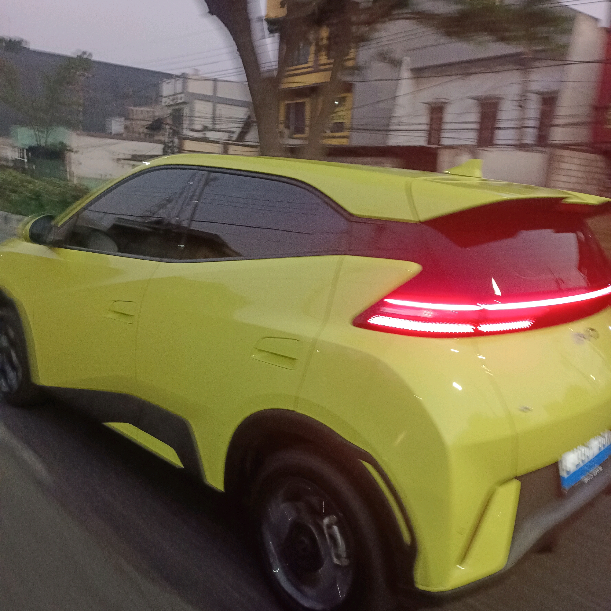 A bright yellow car is captured in motion, its rear end prominently displayed. The car features a distinctive red taillight bar that stretches across the back, with smaller lights underneath. A spoiler extends from the roof, adding to the car's sporty appearance. The car is moving on a paved road, indicated by the motion blur on the road surface. In the background, several buildings are visible, suggesting an urban environment. The sky is overcast and appears to be dusk or dawn. The scene is consistent with a street view in Surabaya, Indonesia.