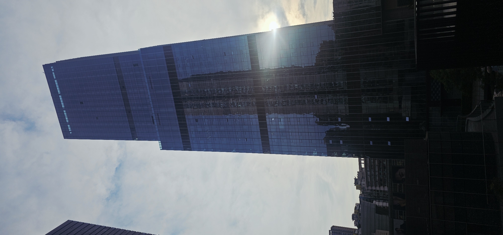 A tall, modern skyscraper with a dark, reflective glass facade dominates the center of the image. The skyscraper is angled, stretching upwards and slightly to the right. Sunlight glints off the glass, creating lens flares and highlighting the surrounding sky, which is partly cloudy and overcast. The reflection on the skyscraper's surface shows other buildings, including a similar-looking, slightly shorter skyscraper. In the foreground and lower portion of the image, there are other buildings with dark facades, some appearing to be commercial or residential structures, and parts of a dark-colored railing or barrier are visible. The overall scene suggests an urban environment, likely in a developed city. This scene is located in Jiulongpo District, China.