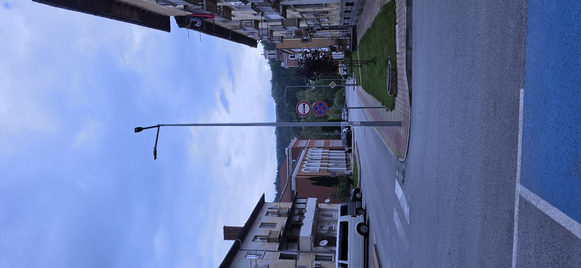The image depicts a street scene in Rudozem, Bulgaria. The street, oriented in a receding perspective, is bordered by buildings of varying heights and architectural styles. A sidewalk is visible on the right side of the street, marked with brick paving. Several parked vehicles, including a white van, are positioned along the right side of the road. Overhead, there is a lamppost extending from the left side of the image. Further down the road, two traffic signs are visible: one indicating "No Passing" and another indicating "No Stopping." In the background, there is a green hillside. The sky is overcast with a mix of light blue and gray clouds. The overall scene suggests a residential area within the city.